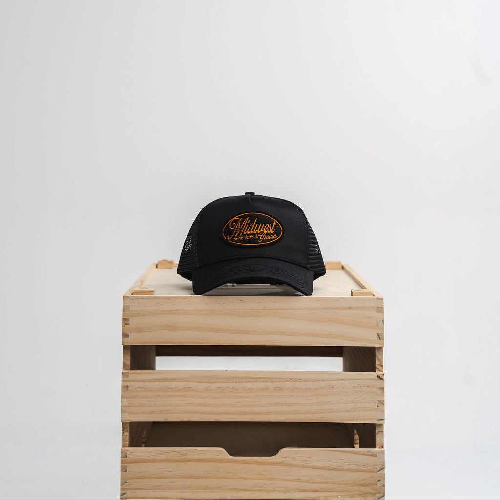 Black cap with gold Midwest Grown logo on a wooden crate against a white background