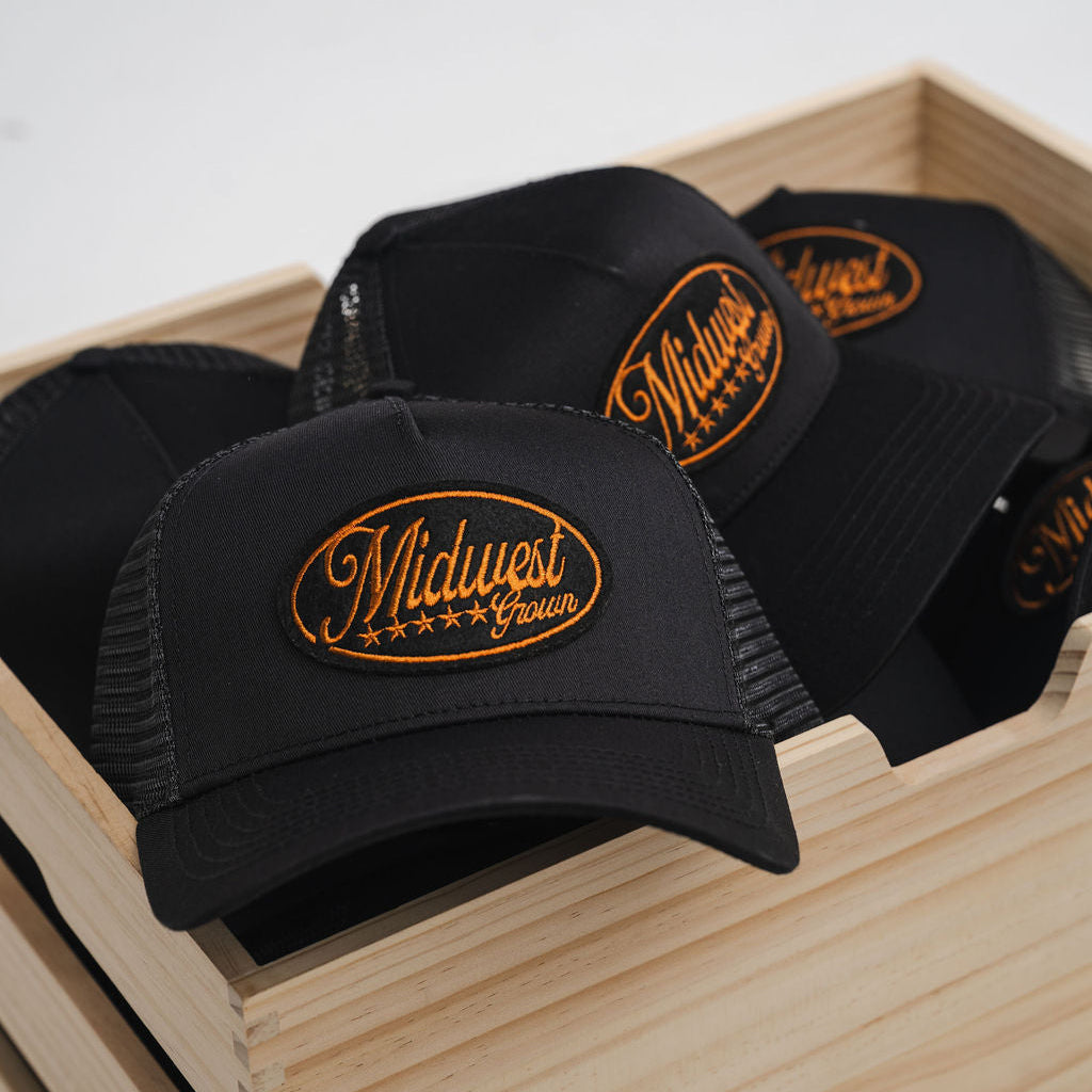Black trucker hats with gold 'Midwest' logo in a wooden box