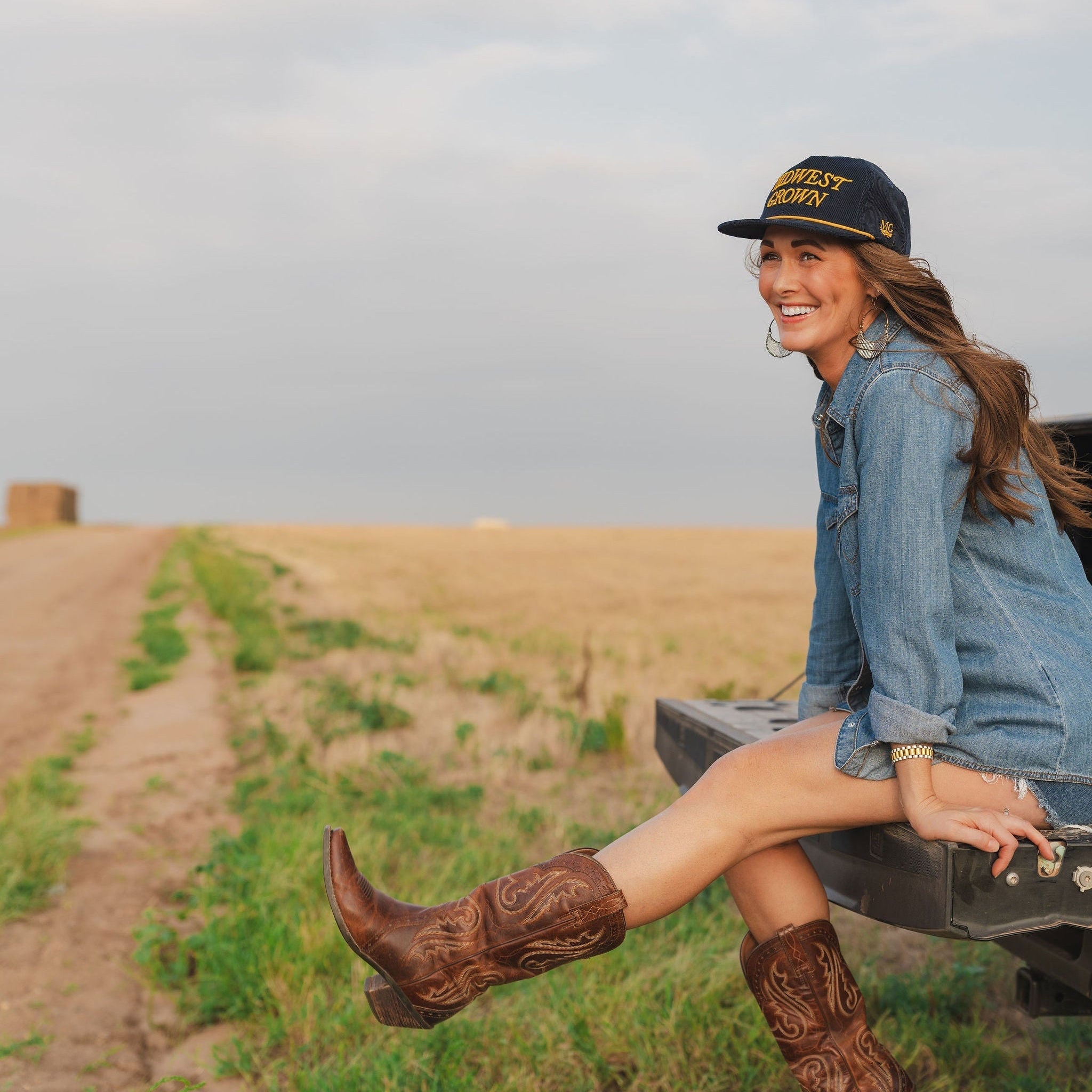 Woman in cowboy boots and denim outfit sitting on a truck bed in a field wearing a Midwest Grown Hat