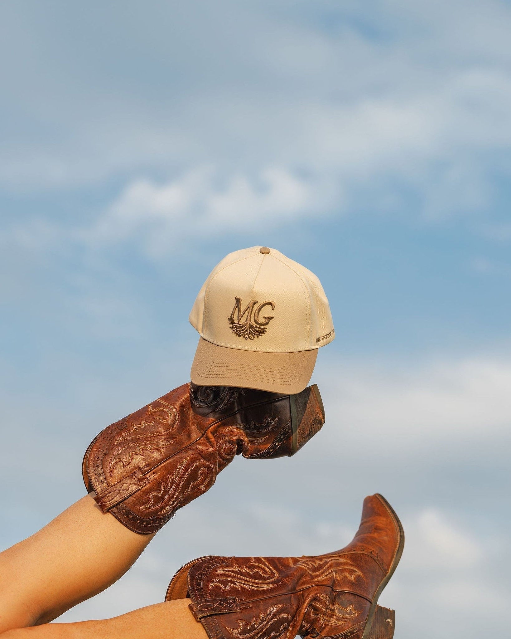 Person wearing cowboy boots and a Midwest Grown cap against a blue sky.