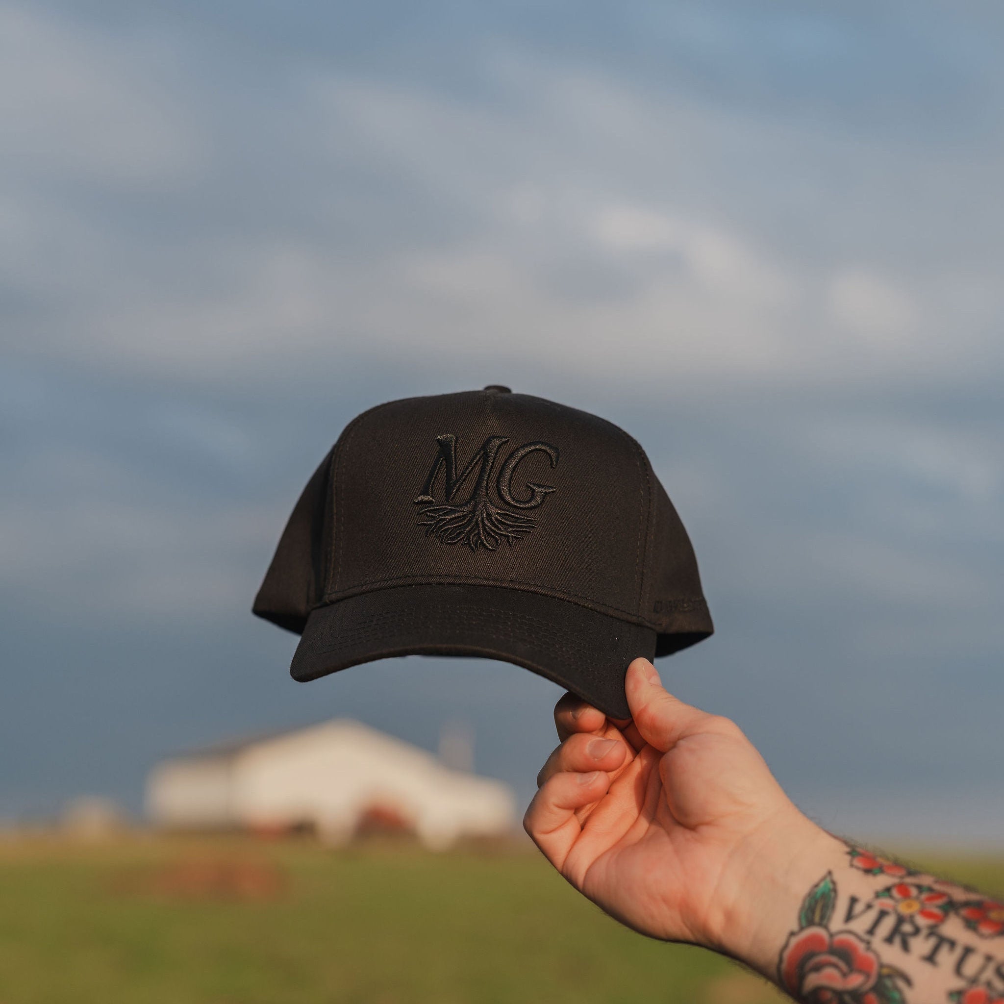 Hand holding a black cap with 'Midwest Grown' logo against a cloudy sky