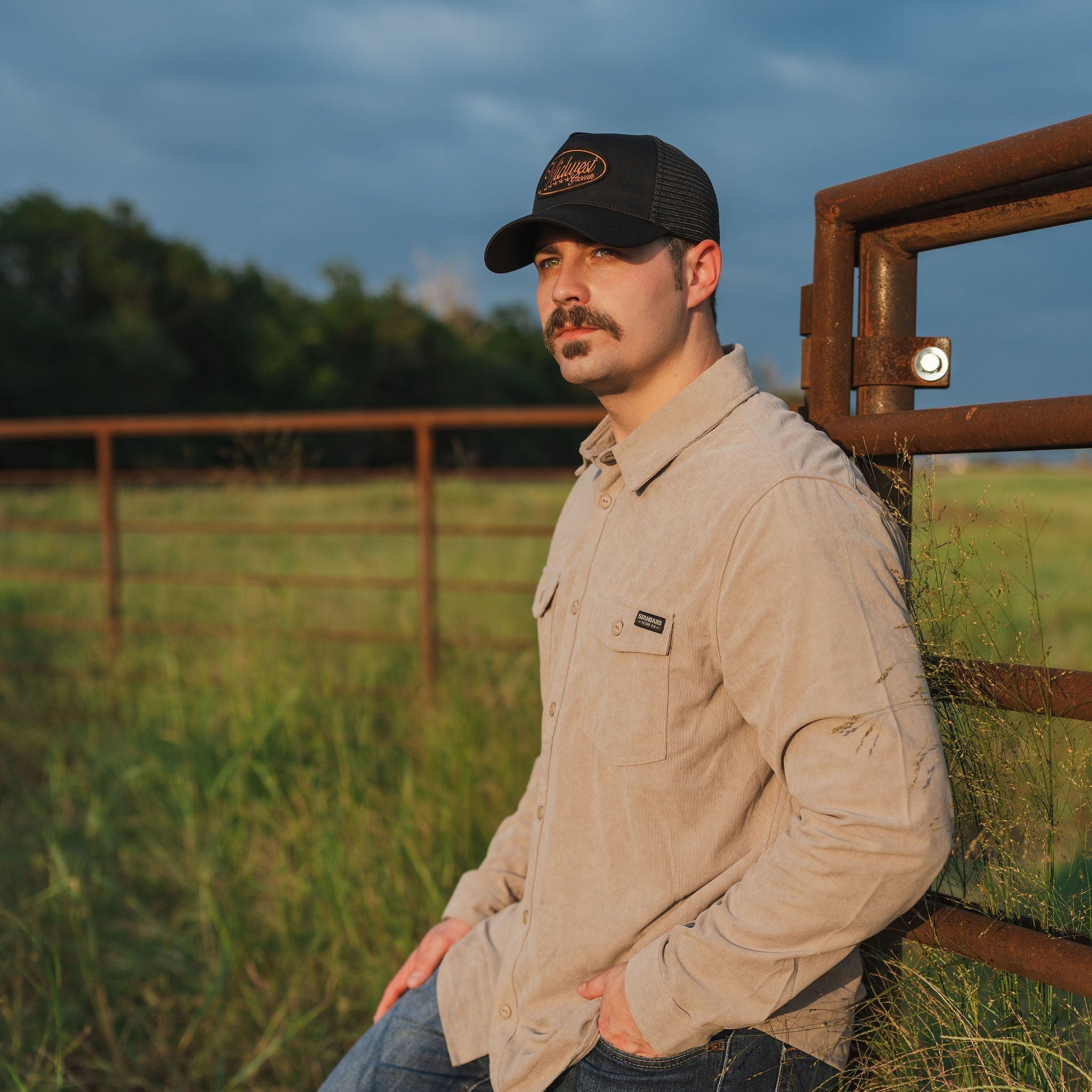 Man leaning against a fence in a field with a dark sky wearing a Midwest Grown hat.