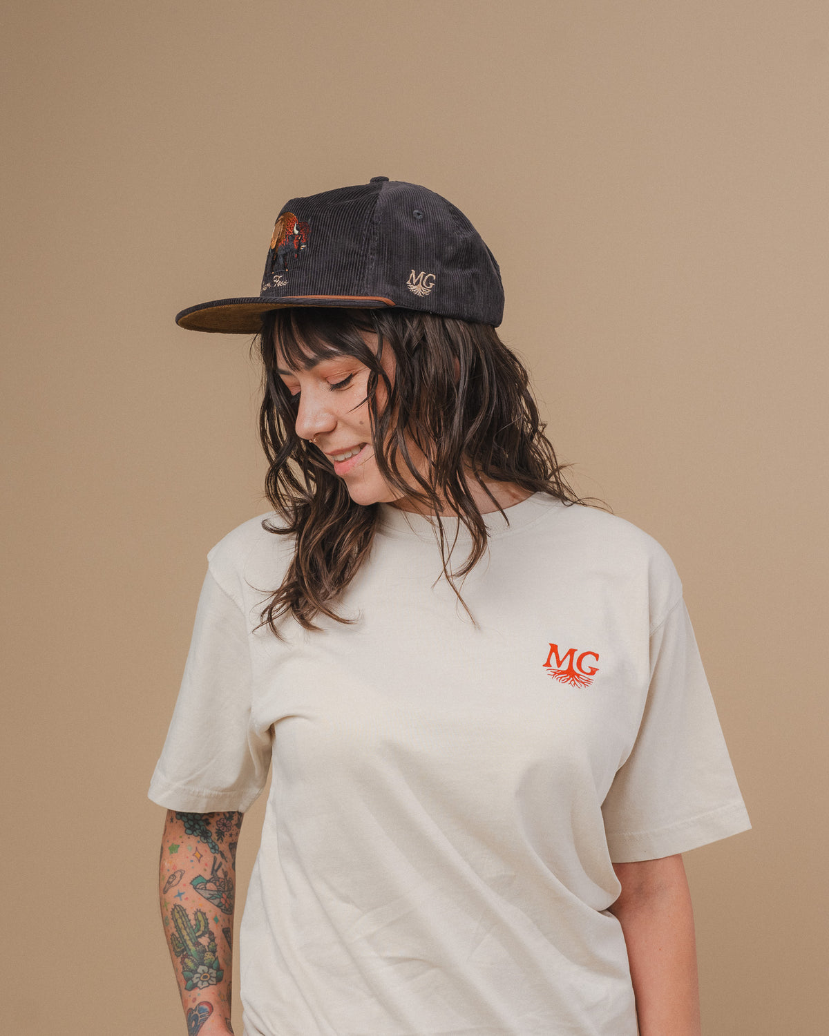 A woman wearing a cream-colored t-shirt with a red 'Midwest Grown' logo on the chest, paired with a dark baseball cap, set against a solid beige background.