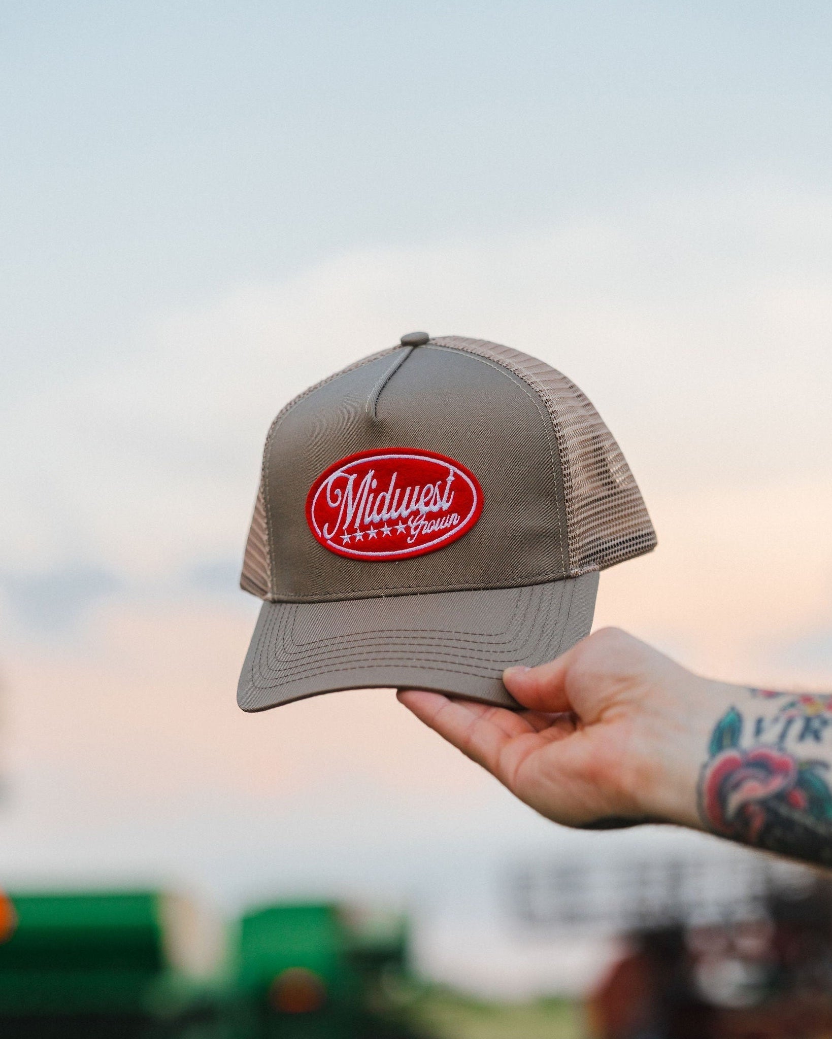 Hand holding a Midwest Grown cap with a logo against a blurred outdoor background
