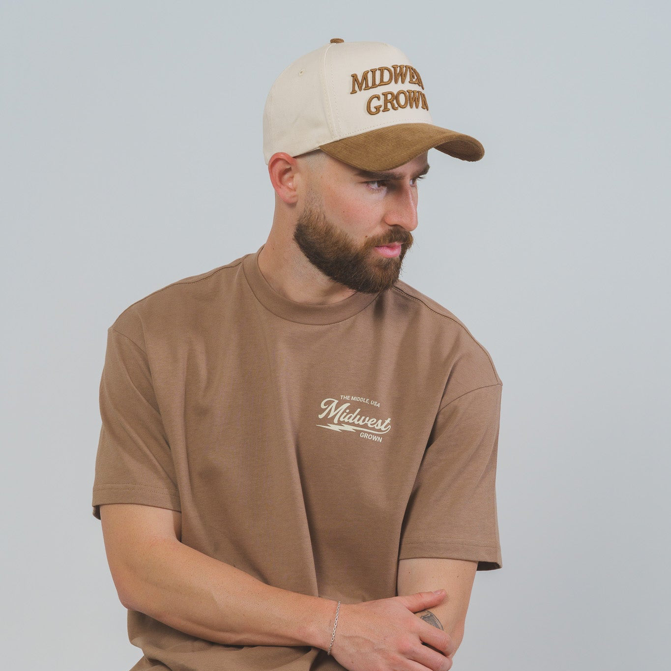 Man wearing a brown t-shirt and cap with 'Midwest Grown' text, sitting on a stool against a plain background.