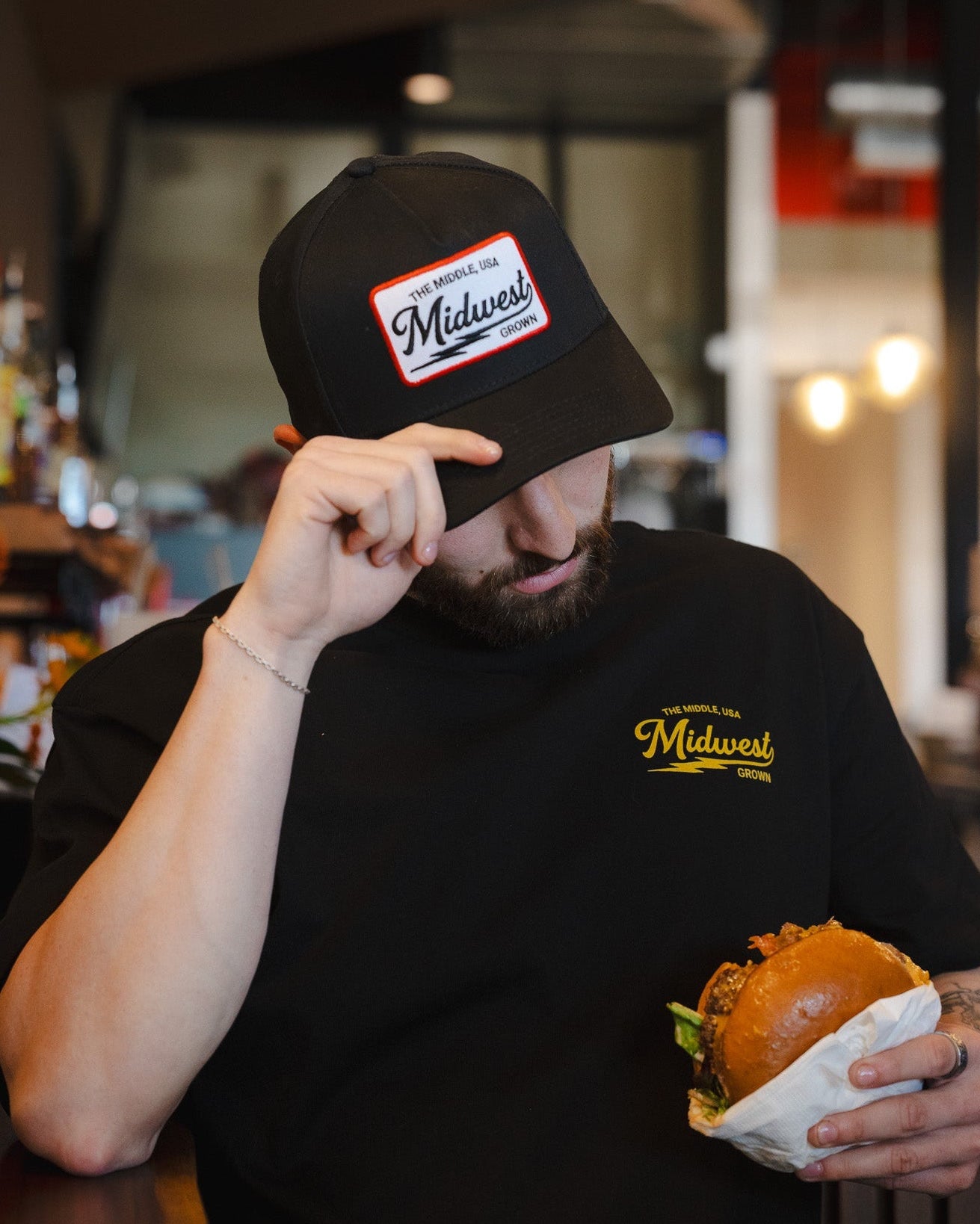Man wearing a black cap and t-shirt with 'Midwest' logo, holding a burger in a casual setting.