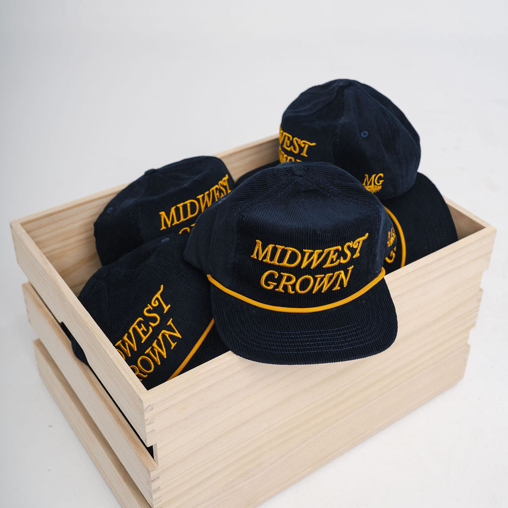 Navy colored hats with 'Midwest Grown' text in yellow on a wooden crate against a white background