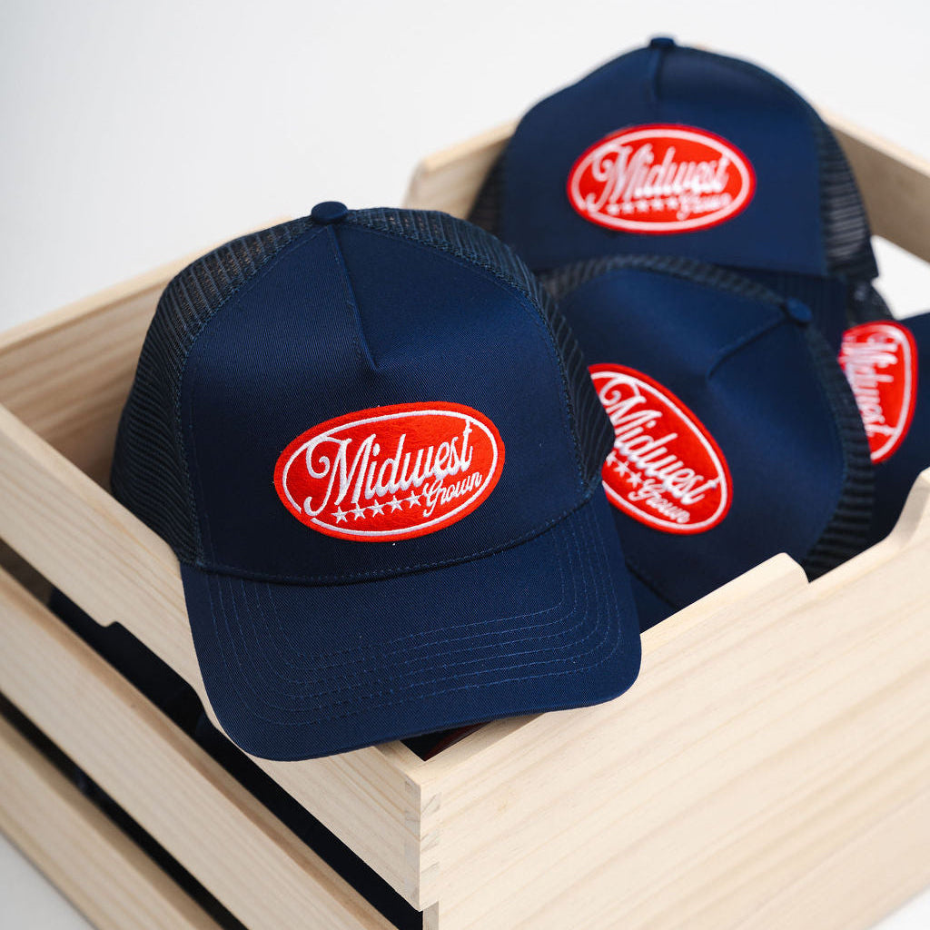 Navy blue caps with red logo reading Midwest Grown in a wooden box on a white background