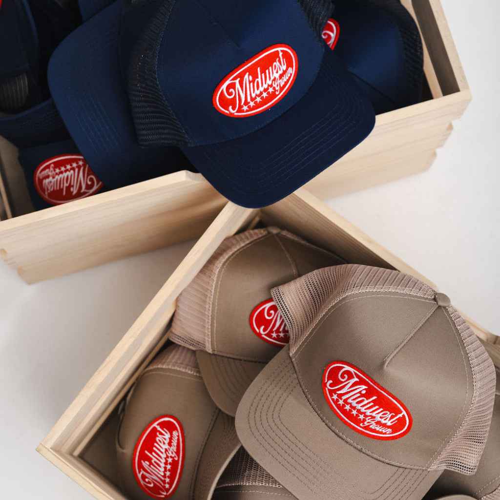 Navy and brown Midwets Grown caps with a red logo in wooden boxes on a white surface