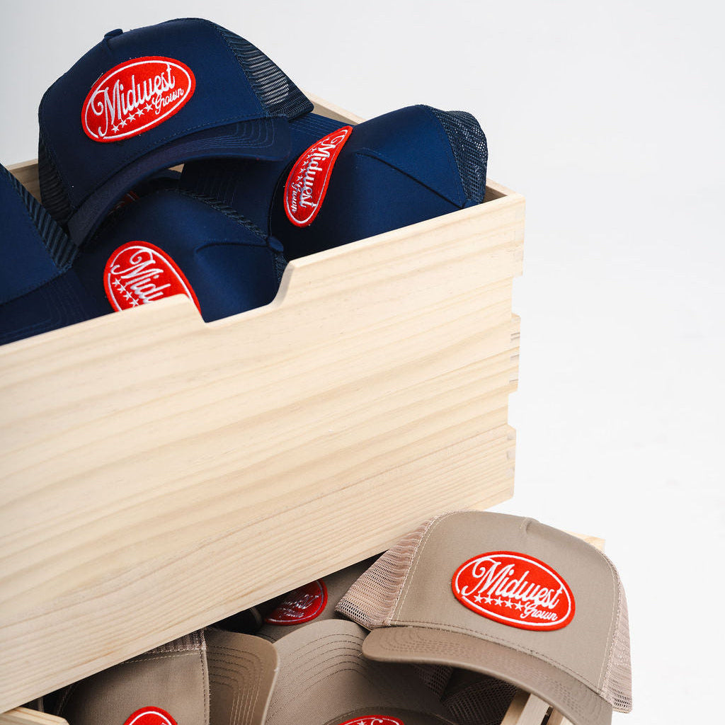 Navy and beige Midwest Grown baseball caps with a red logo in a wooden box on a light gray background