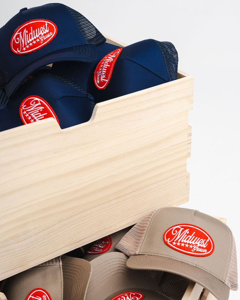 Navy and beige Midwest Grown baseball caps with a red logo in a wooden box on a light gray background