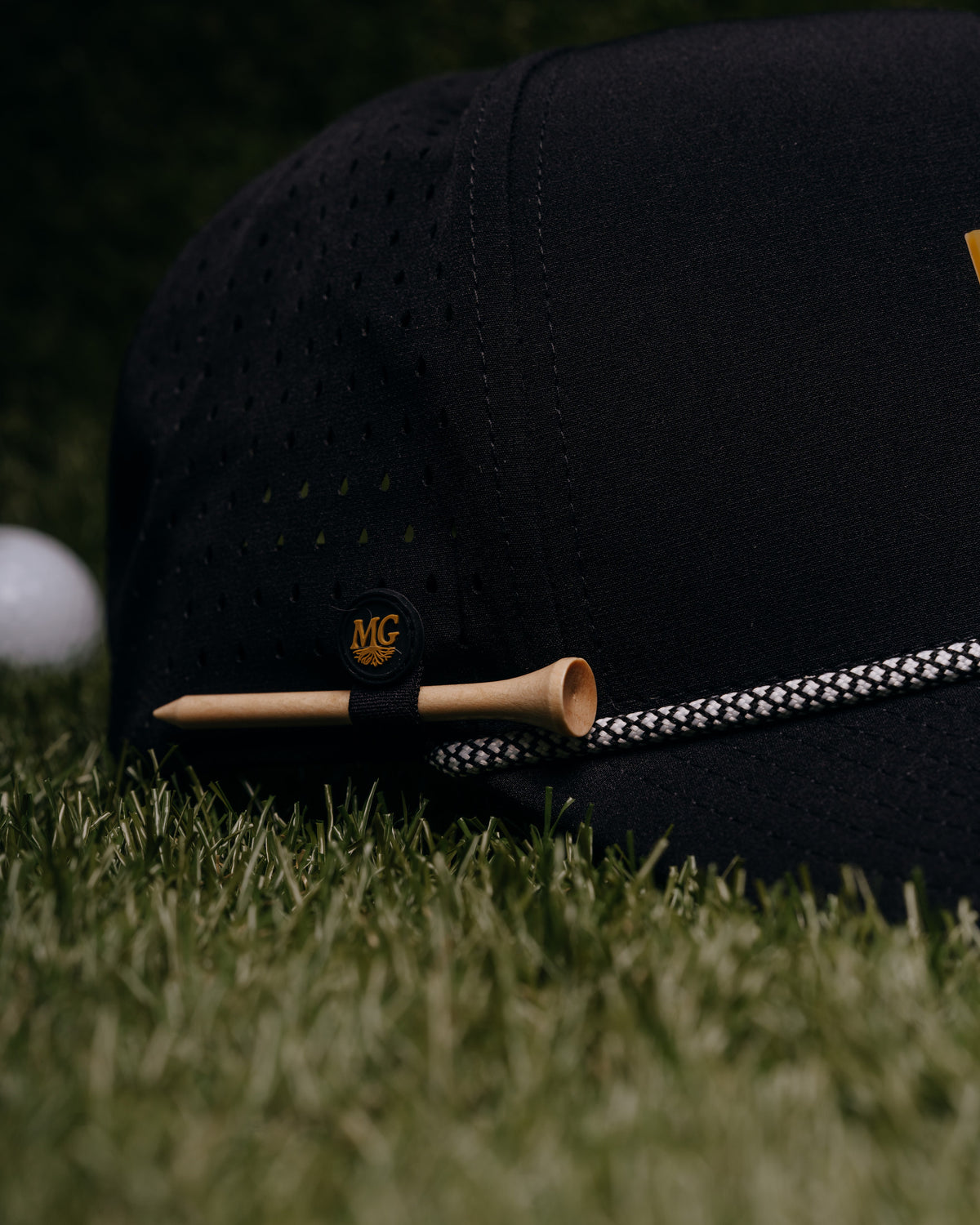 A black Midwest Grown perforated baseball cap resting on green grass, with a wooden tee and a white golf ball nearby. The cap features a small black patch with a gold monogram logo and a black-and-white patterned rope detail.
