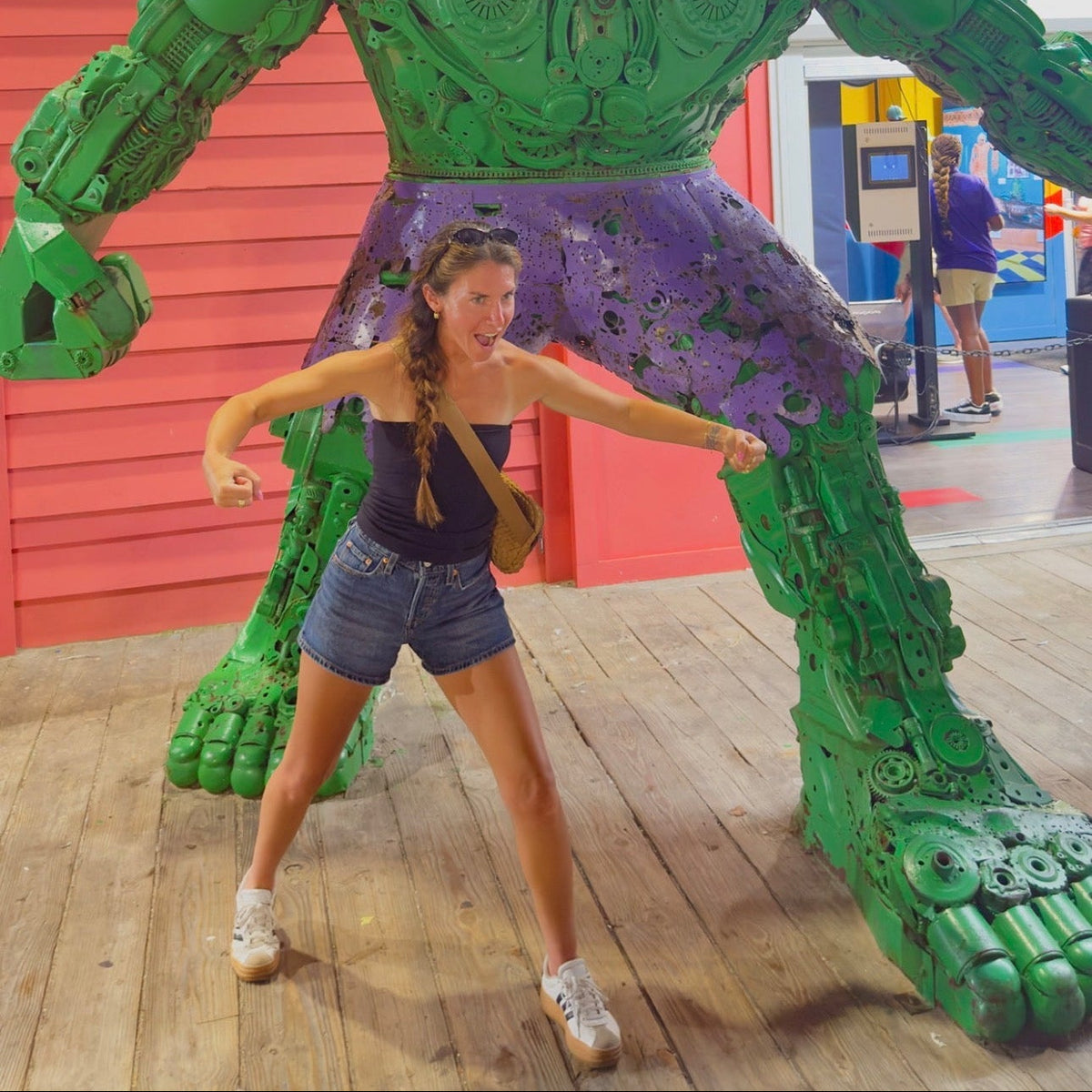 A woman posing in front of a giant green metal sculpture resembling the Hulk, set against a red corrugated metal wall and wooden floor.