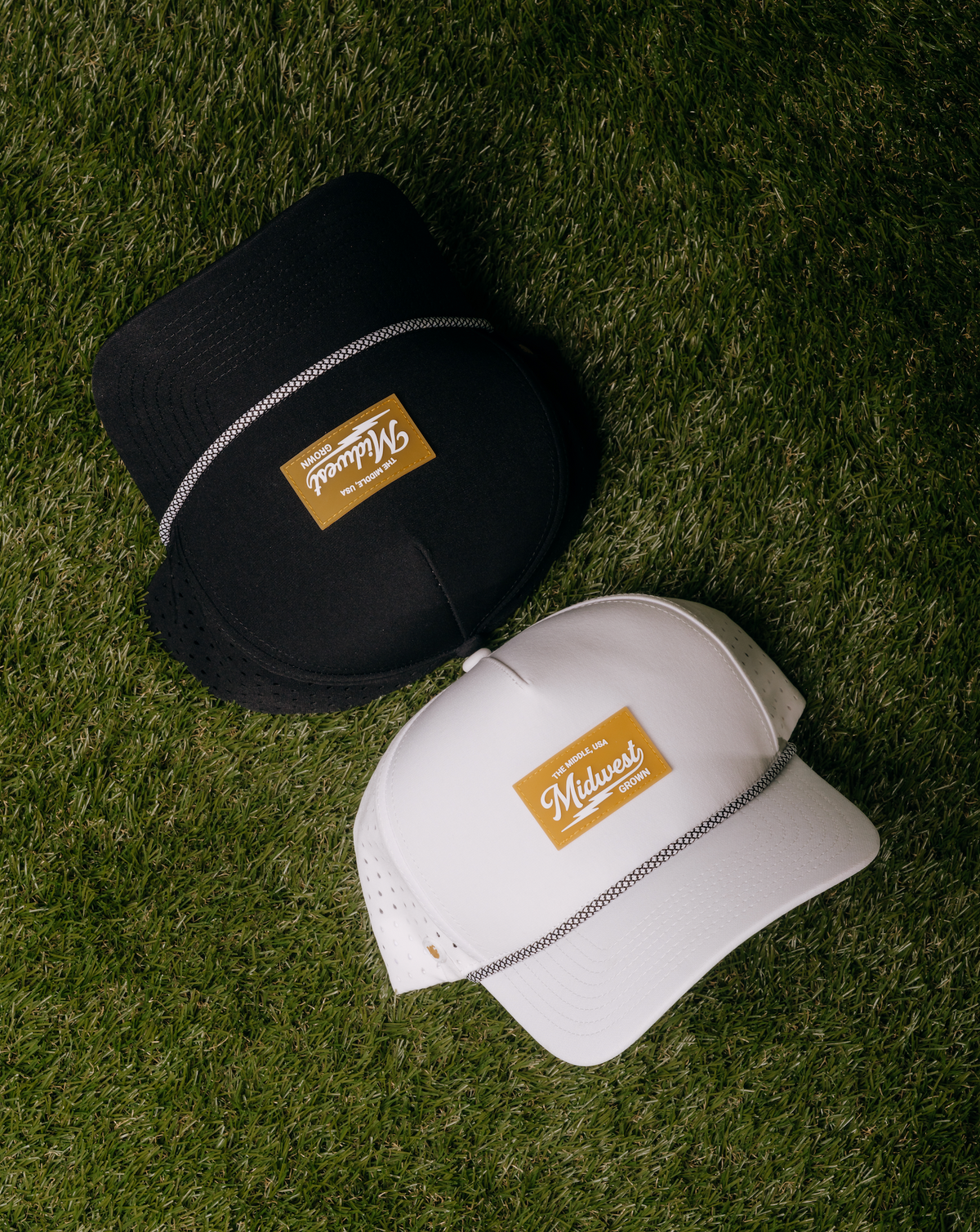 Two baseball caps with a gold 'Midwest' patch and silver chain detail, displayed on green grass. One cap is black and the other is white.