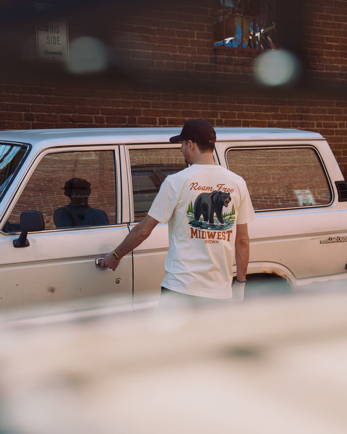 A man wearing a white t-shirt with a large Midwest bear graphic stands next to a vintage white station wagon in front of a brick wall.