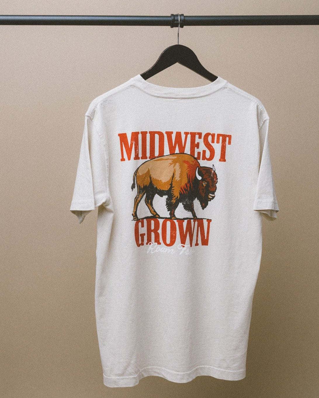 White t-shirt with a graphic of a bison and 'Midwest Grown' text on a hanger against a beige background