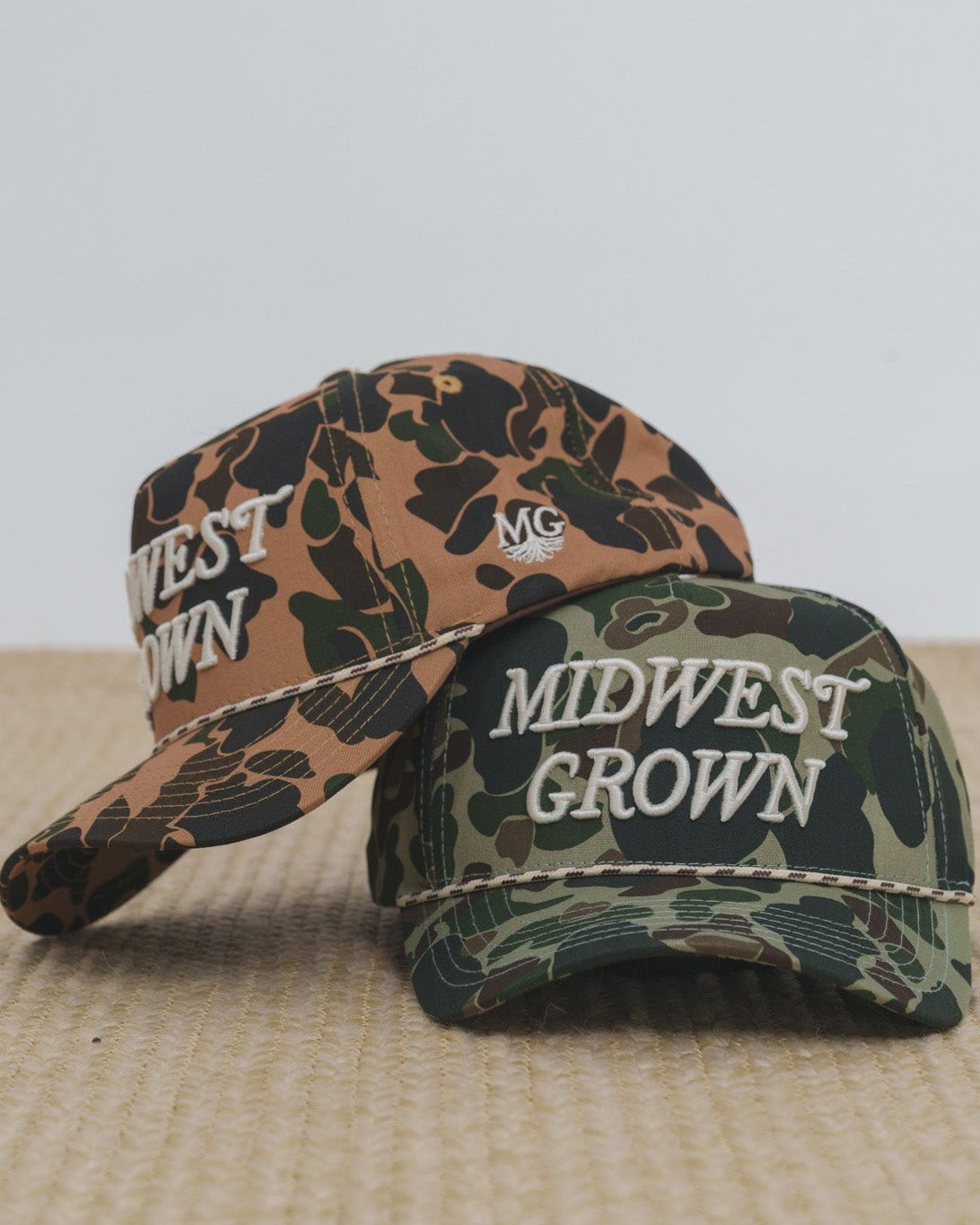 Two duck camo baseball caps with 'Midwest Grown' text on a textured surface.