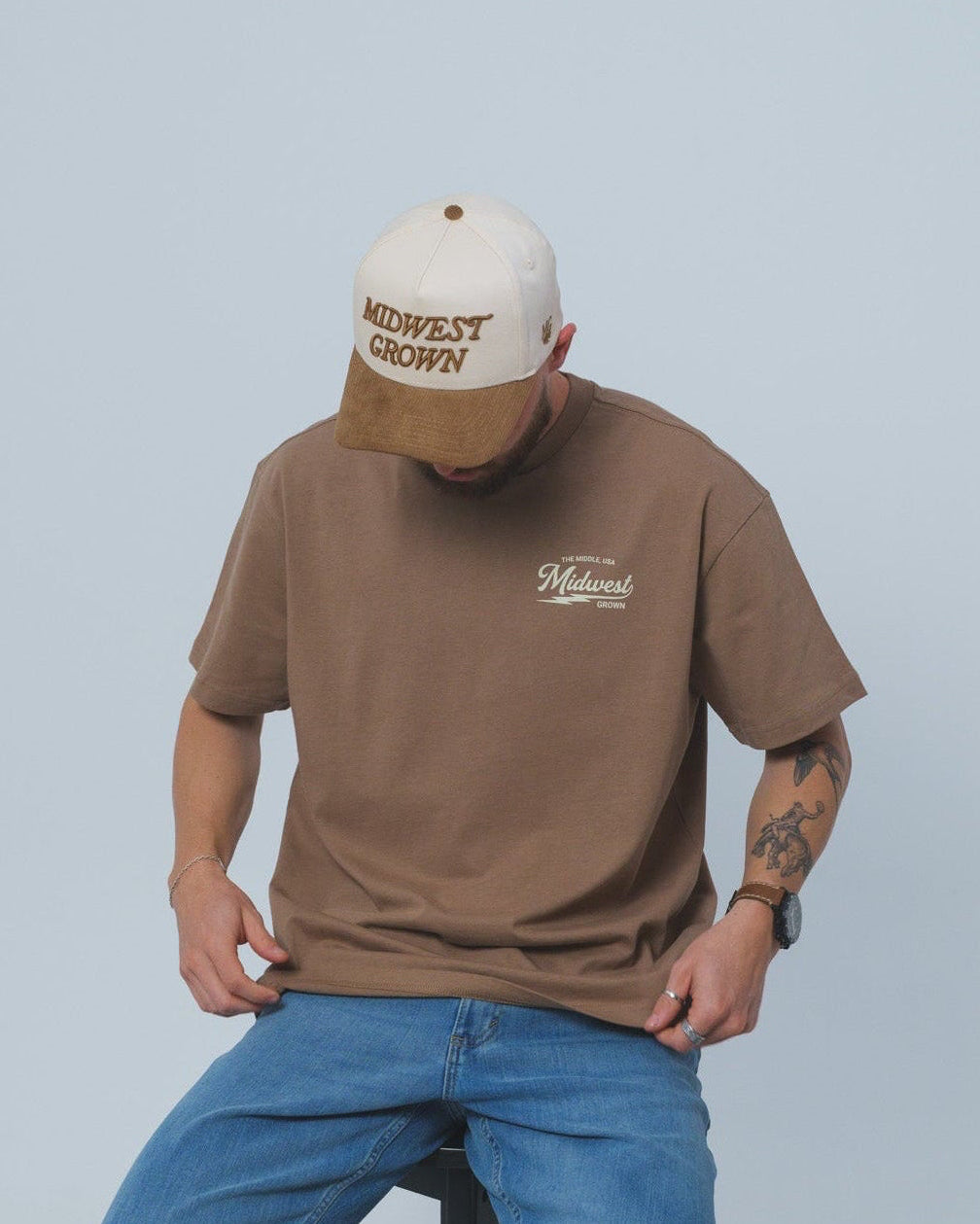 Man wearing a brown Midwest Grown shirt and blue jeans sitting on a stool against a plain background