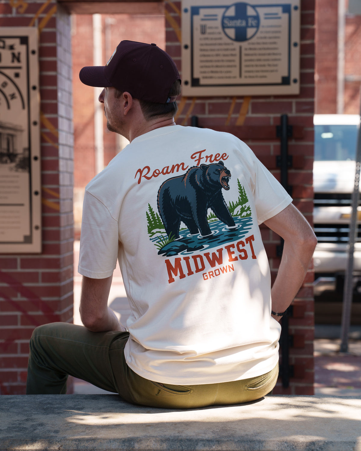 A man wearing a cream-colored t-shirt with a large back graphic of a black bear and the text 'Roam Free Midwest Grown', sitting outdoors in front of a brick wall.