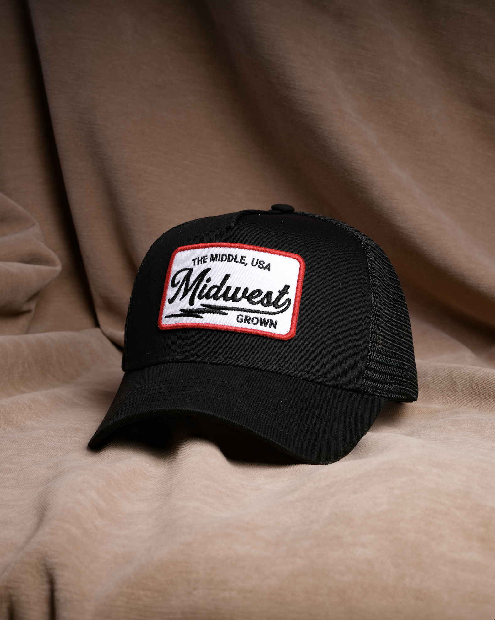 A black trucker-style cap with a white rectangular front patch reading 'The Middle, USA Midwest Grown' in black script and border, photographed against draped tan fabric.