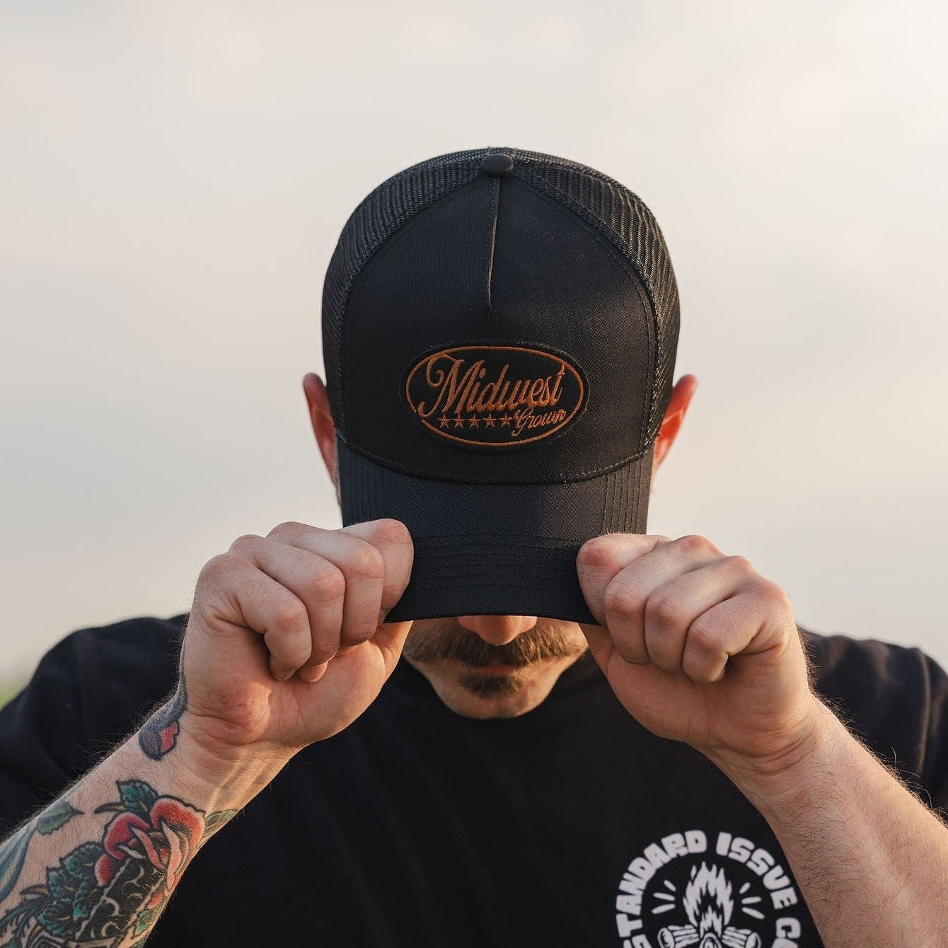 Person holding a black cap with 'Midwest Grown' logo in front of their face, wearing a black t-shirt with a logo, against a blurred natural background.