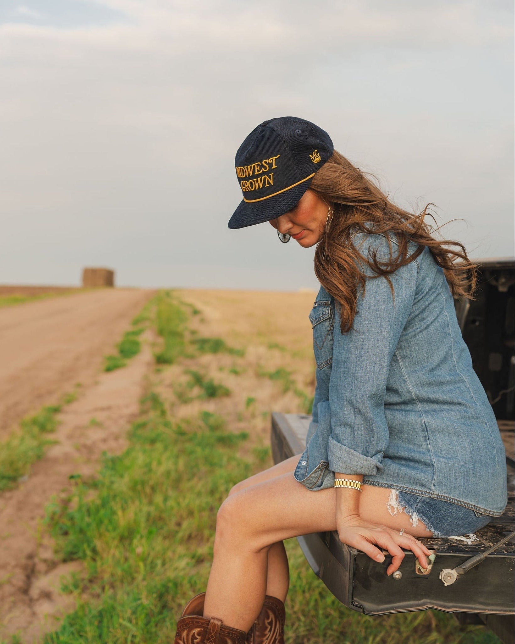 Woman sitting on a truck bed in a field wearing cowboy boots, a denim jacket, and a Midwest Grown Hat.