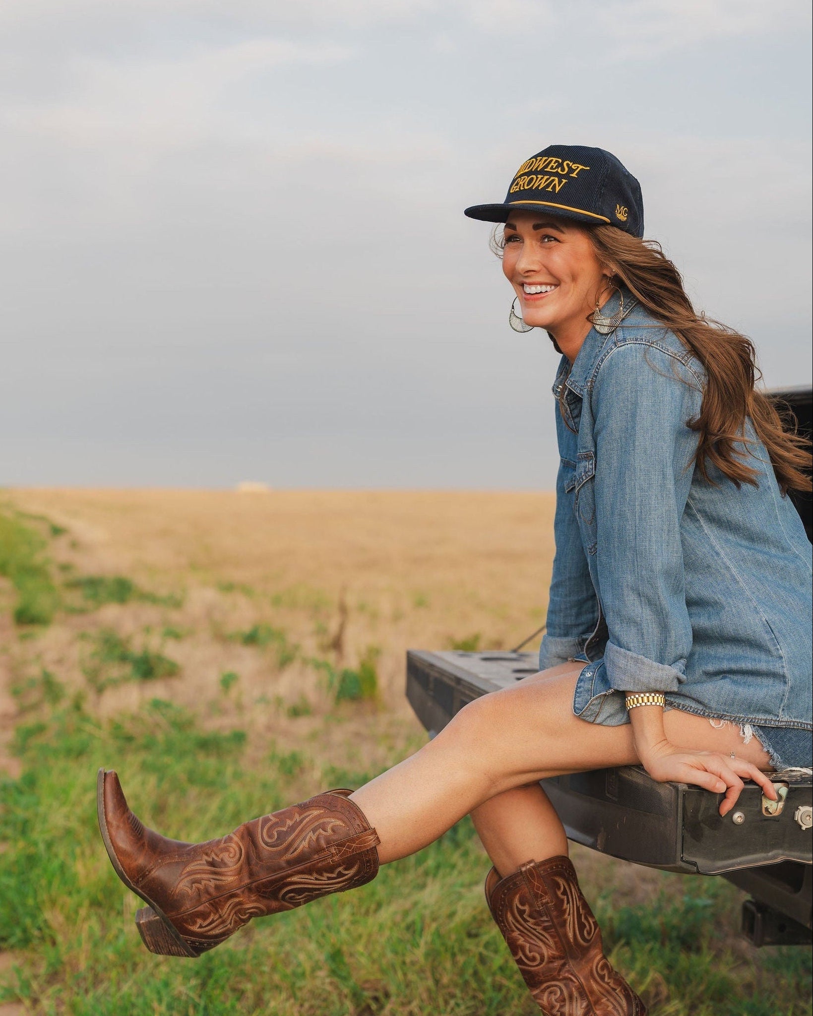 Woman in cowboy boots and denim outfit sitting on a truck bed in a field wearing a Midwest Grown Hat
