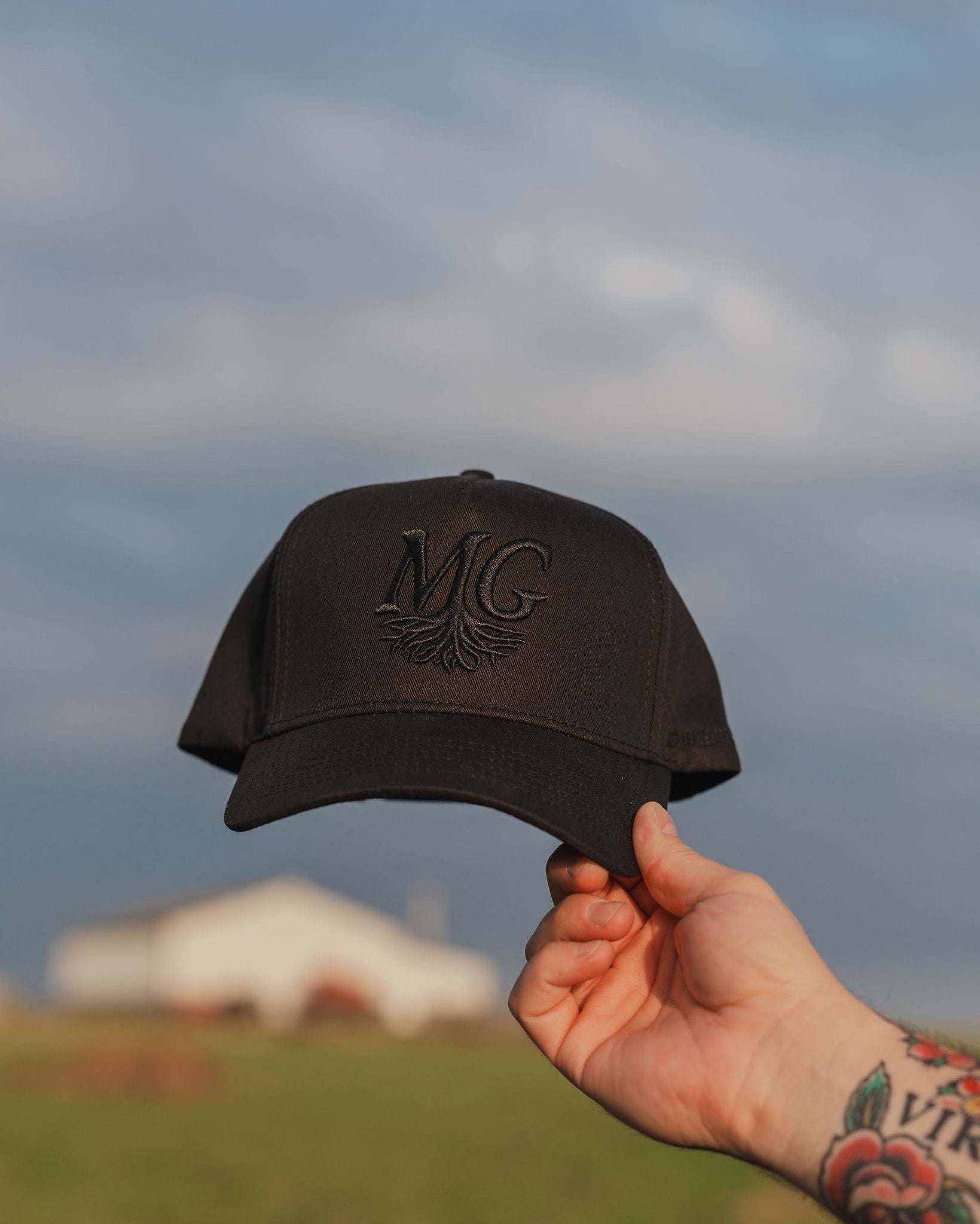 Hand holding a black cap with 'Midwest Grown' logo against a cloudy sky