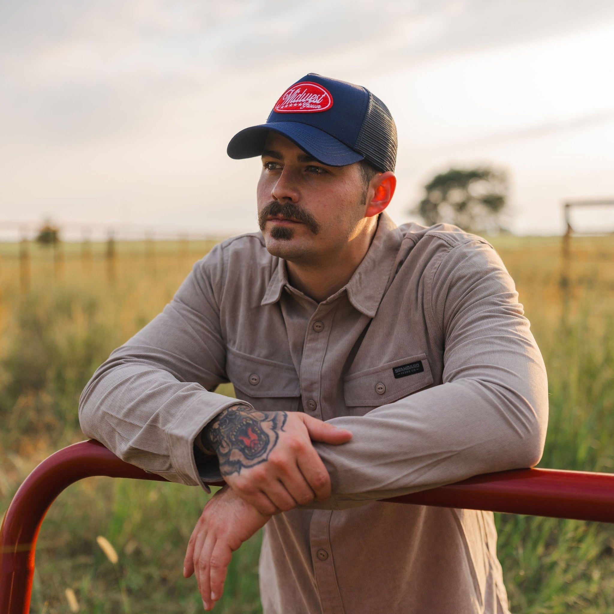Man wearing a Midwest Grown cap and leaning on a red metal gate in a field