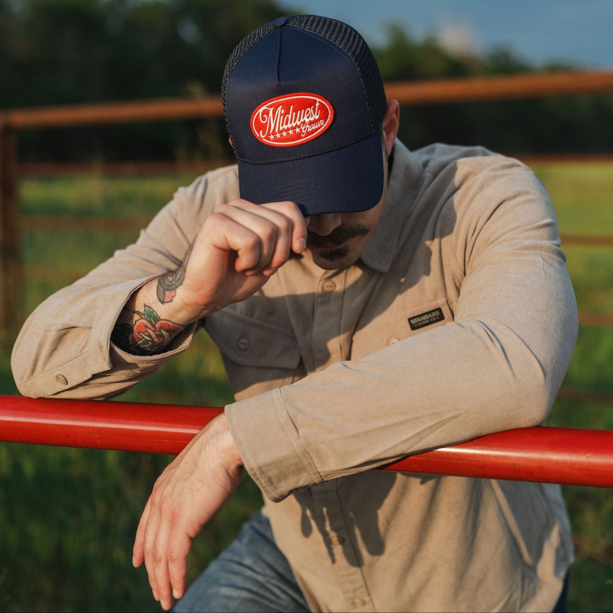 Man wearing a navy cap with a red logo that reads Midwest Grown, sitting on a red fence in an outdoor setting.