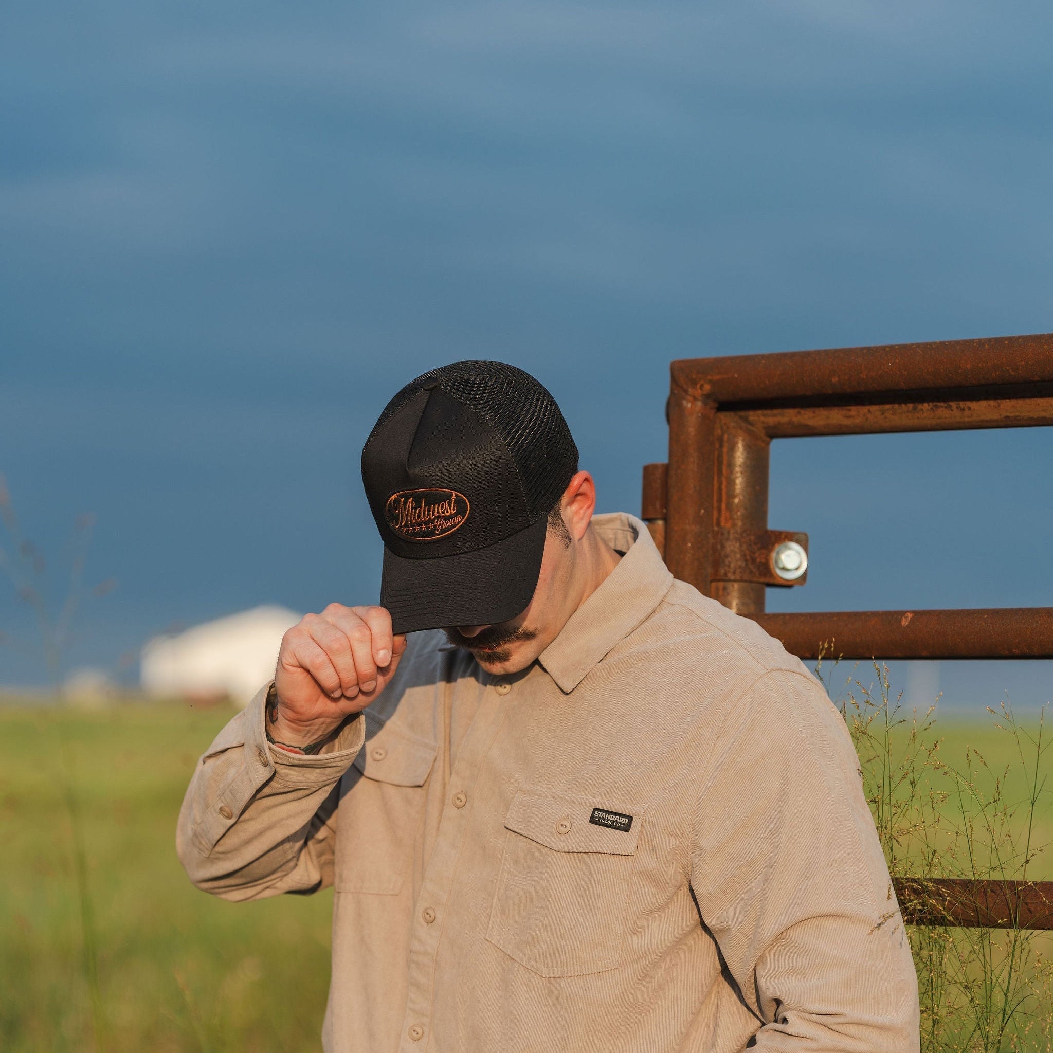 Man wearing a beige jacket and black Midwest Grown  cap in a grassy field with a fence and blue sky.
