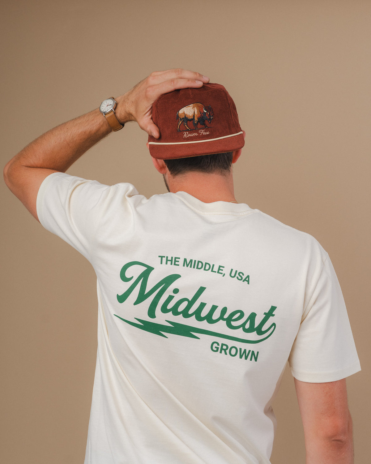 Person wearing a white t-shirt with 'The Middle, USA Midwest Grown' text and a brown cap with a logo.