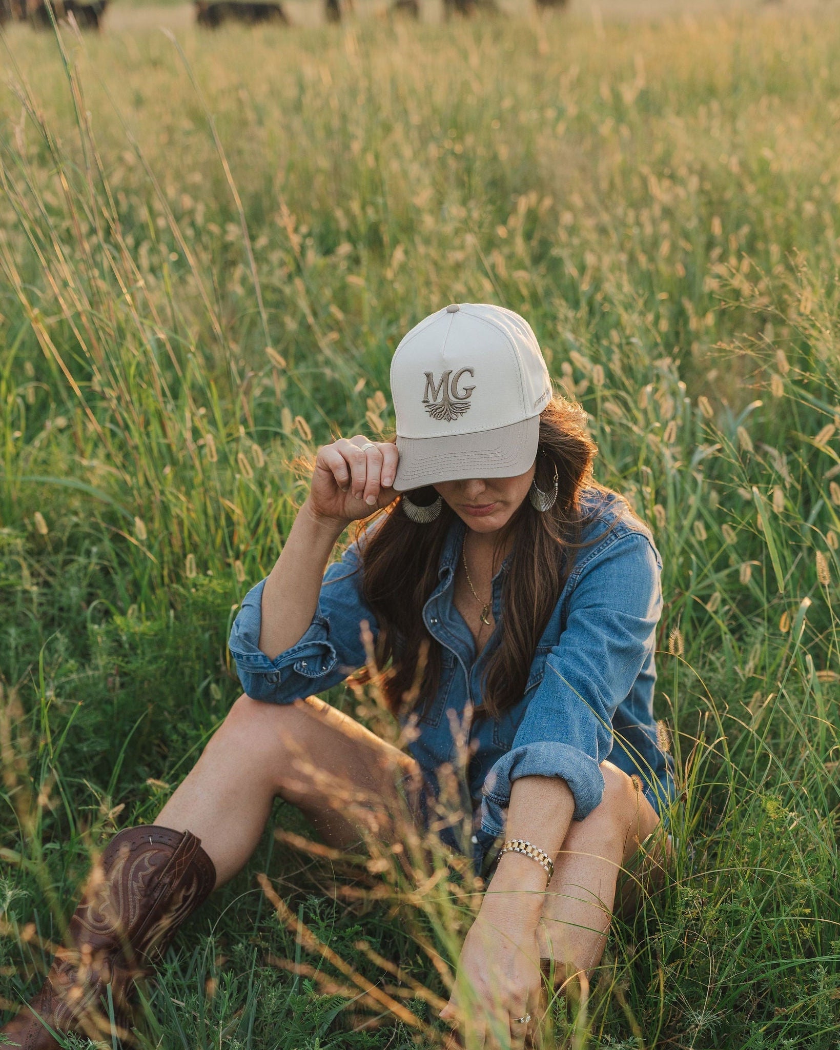 Person sitting in a field wearing a denim jacket and cream Midwest Grown cap.