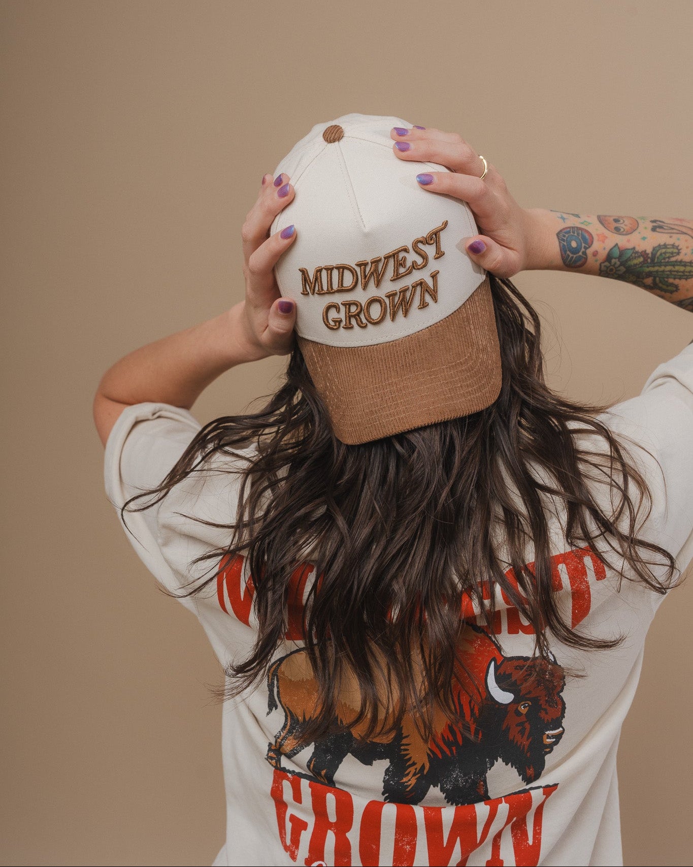 Person wearing a white t-shirt with 'Midwest Grown' design, holding a cap with the same text against a beige background.