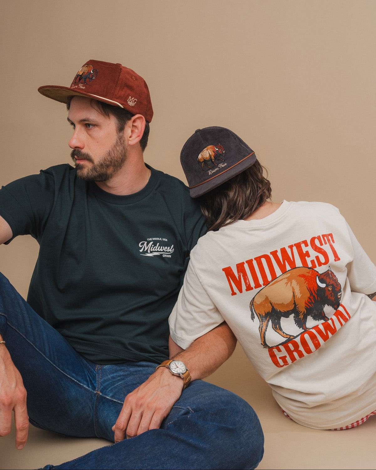 Two models sitting on a beige floor against a beige wall, wearing Midwest Apparel products. The man on the left wears a dark green t-shirt and a brown cap, while the woman on the right wears a white t-shirt with a large buffalo graphic and 'MIDWEST GROWN' text, and a dark cap.