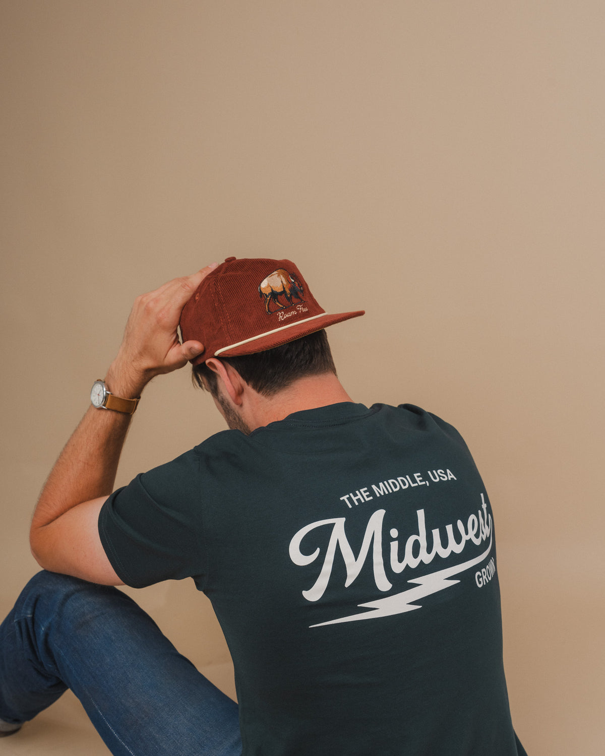 A man sitting on the floor, seen from behind, wearing a dark green t-shirt with 'Midwest' printed in white on the back and a rust-brown baseball cap with a bison graphic.