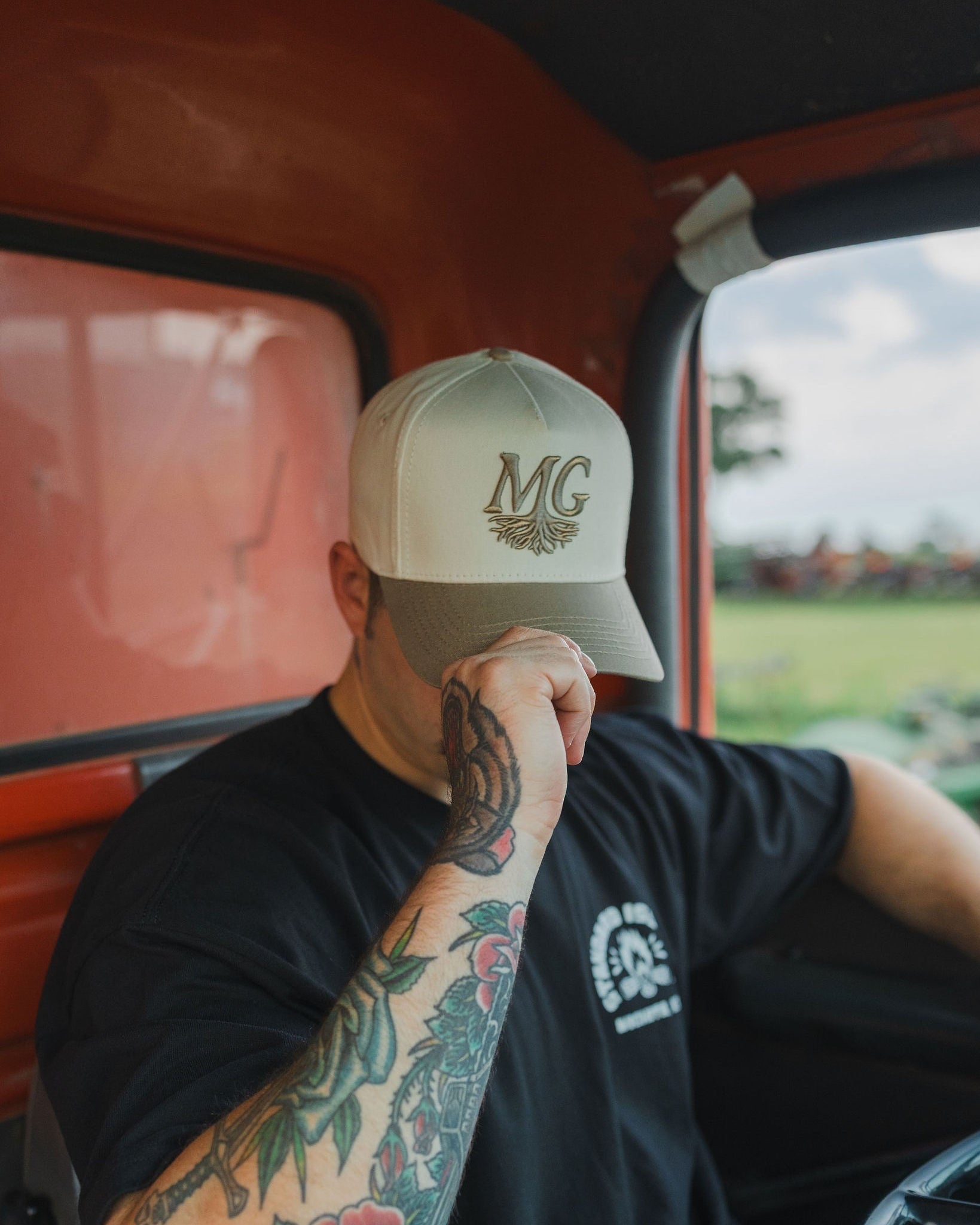 Person sitting inside a vehicle wearing a cap with 'Midwest Grown' logo, tattoos on arm, and a black t-shirt.