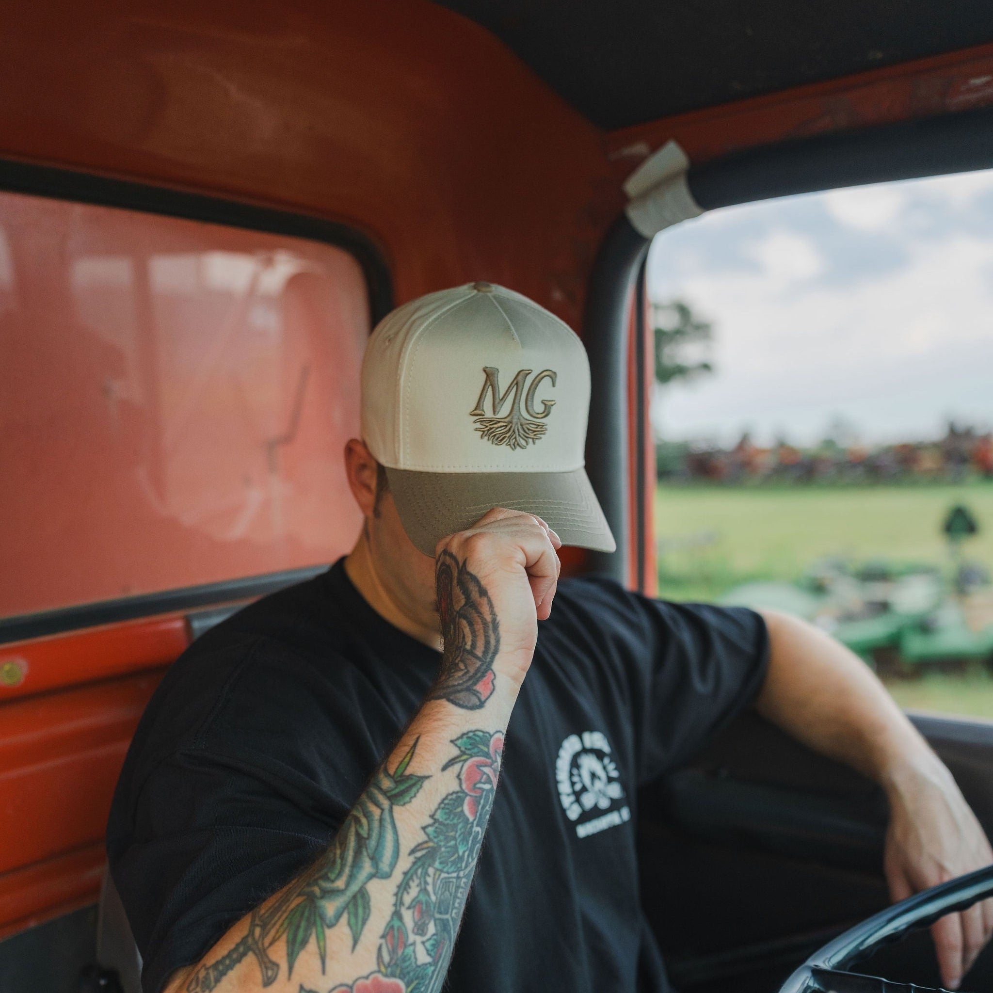 Person sitting inside a vehicle wearing a cap with 'Midwest Grown' logo, tattoos on arm, and a black t-shirt.