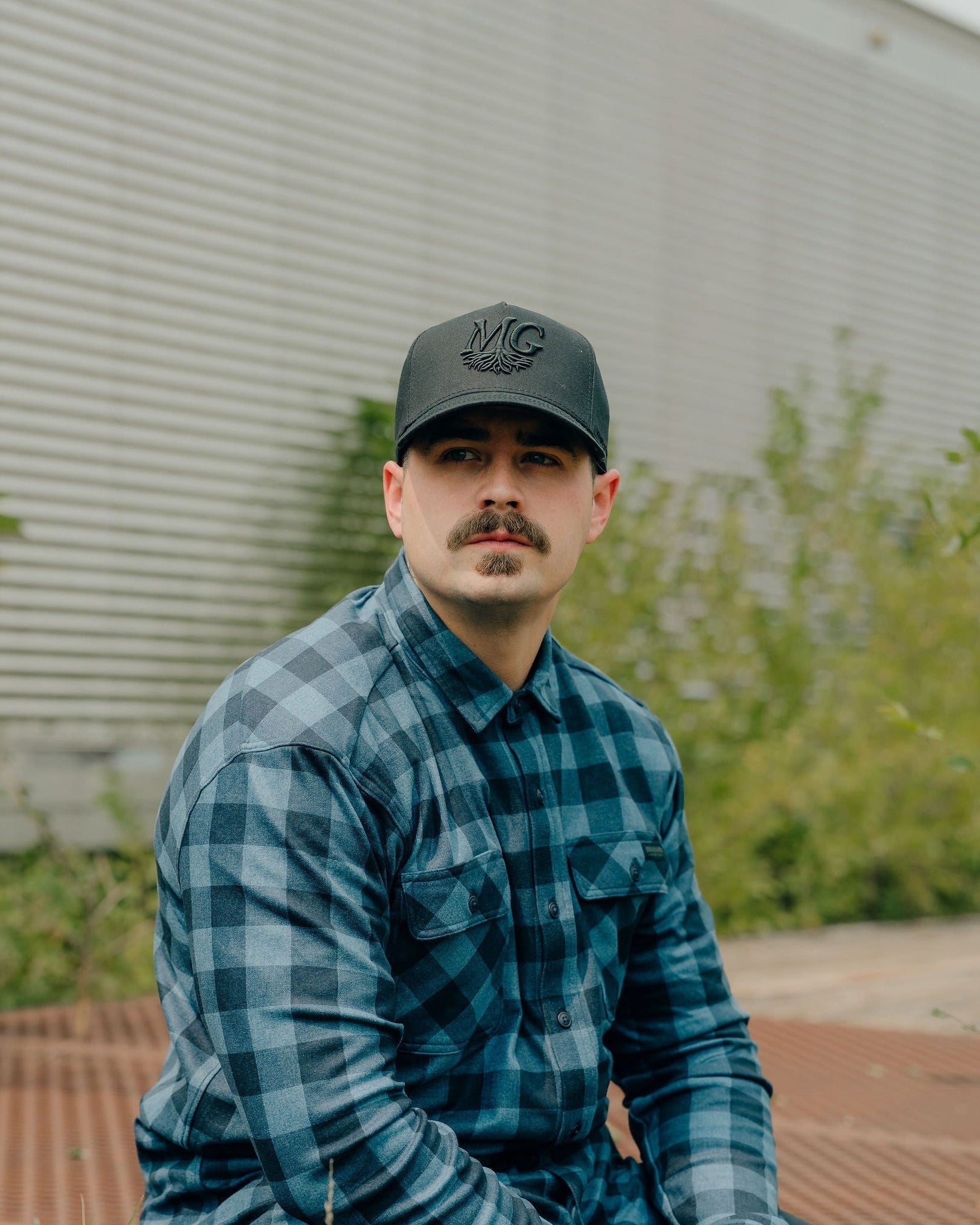 Man wearing a blue plaid shirt and a Midwest Grown cap sitting on a metal surface with greenery in the background