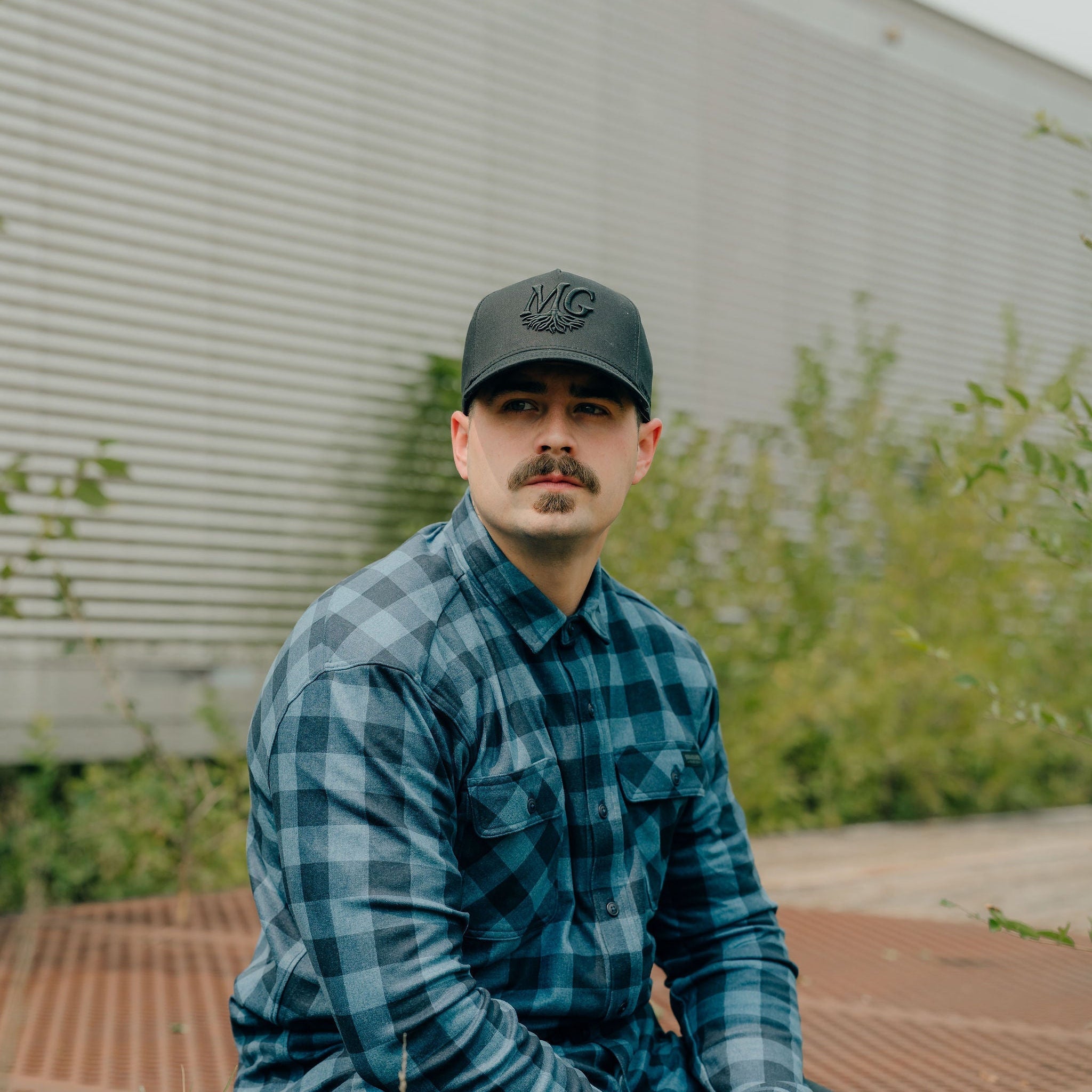 Man wearing a blue plaid shirt and a Midwest Grown cap sitting on a metal surface with greenery in the background