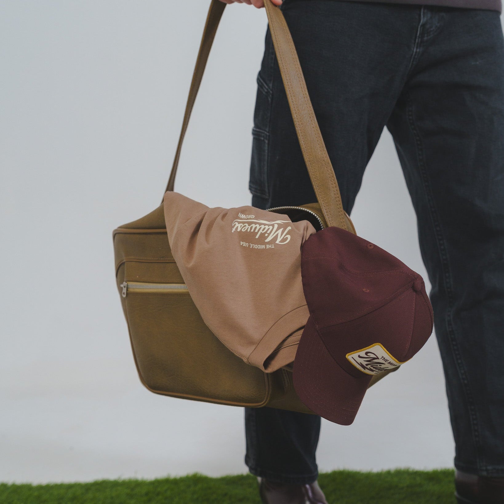 Person holding a brown bag with a maroon Midwest Grown cap on a grassy surface