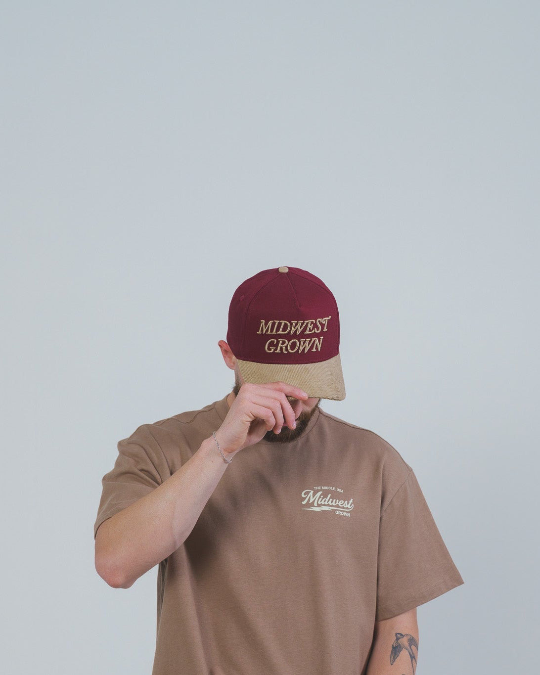 Person wearing a brown t-shirt and blue jeans with a maroon and beige cap on a plain background