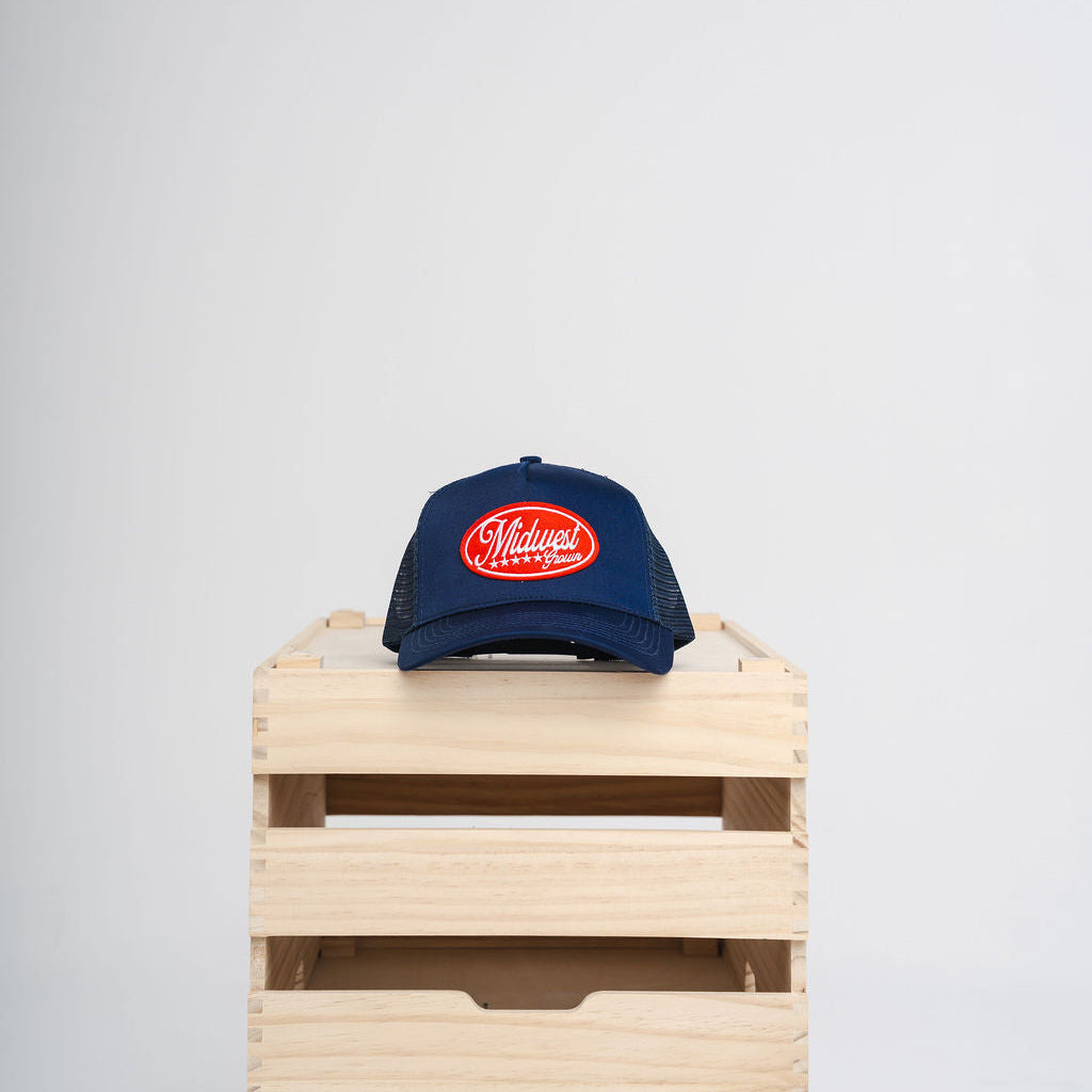 Blue Midwest Grown cap with a red logo on a wooden crate against a white background