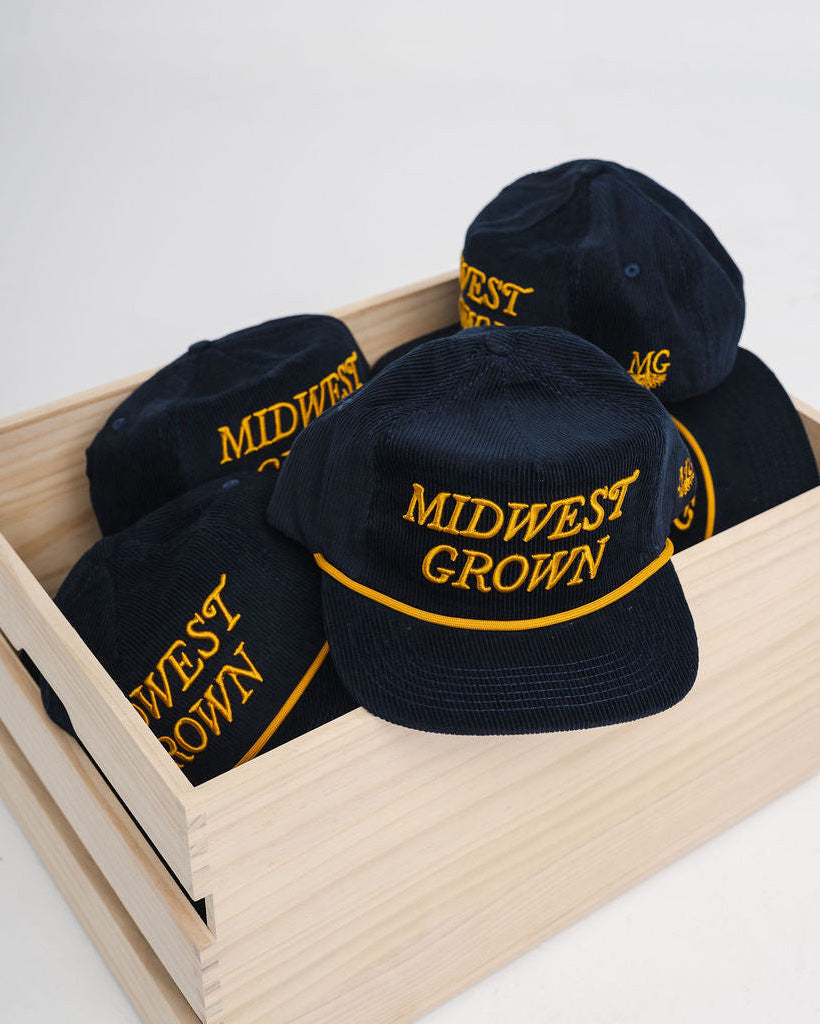Navy colored hats with 'Midwest Grown' text in yellow on a wooden crate against a white background