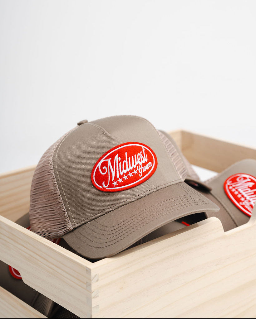 Beige Midwest Grown cap with red logo in a wooden crate on a light gray background