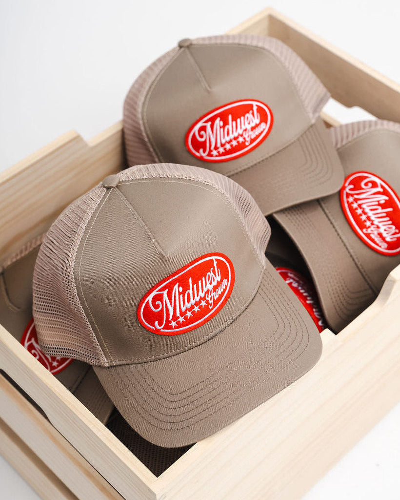 Three beige Midwest Grown caps with red logos in a wooden box on a light gray background