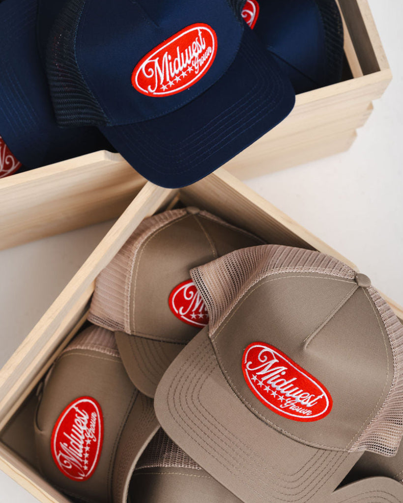 Navy and brown Midwets Grown caps with a red logo in wooden boxes on a white surface