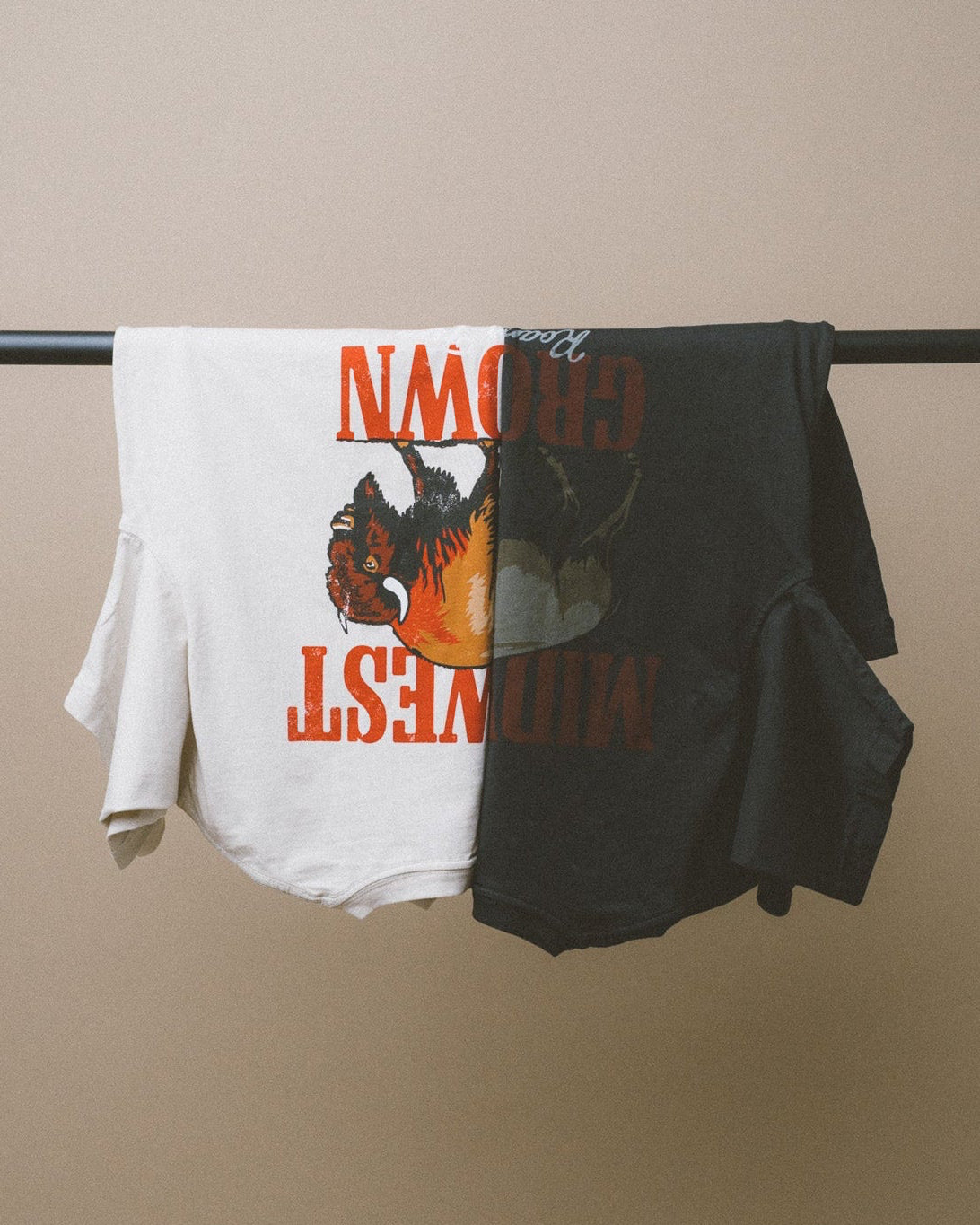 Two Midwest Grown t-shirts, one white and one black, with graphic designs and text, hanging on a rod against a beige wall.