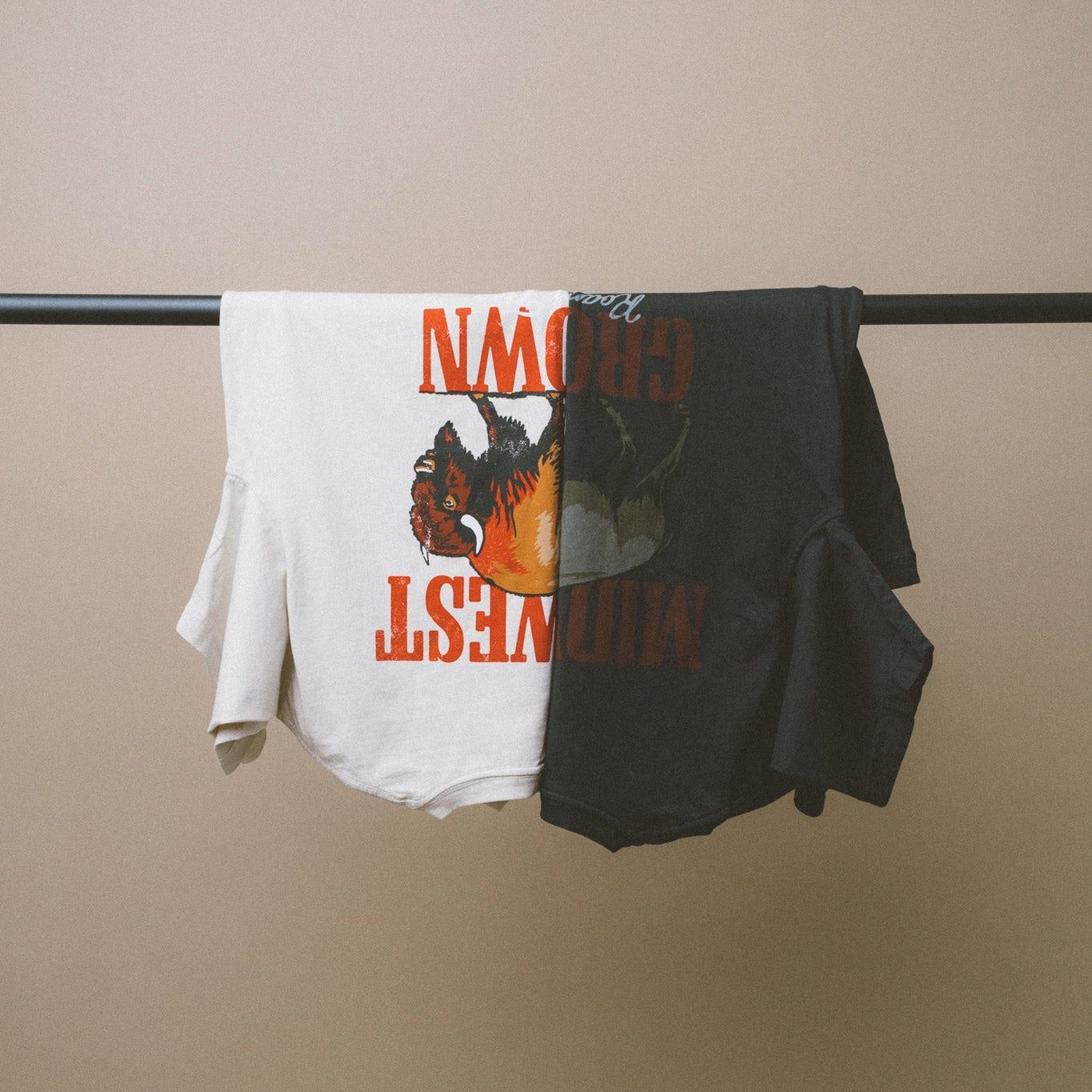 Two Midwest Grown t-shirts, one white and one black, with graphic designs and text, hanging on a rod against a beige wall.
