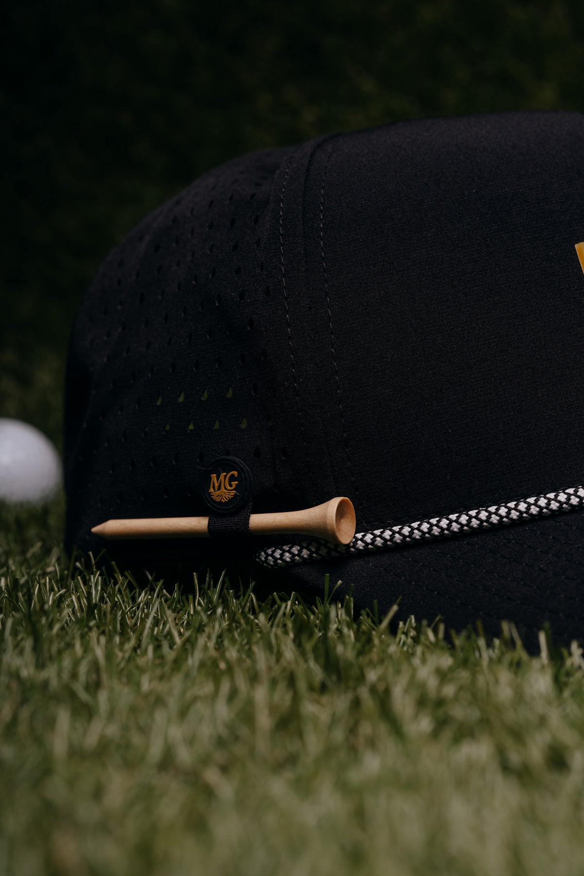 A black Midwest Grown perforated baseball cap resting on green grass, with a wooden tee and a white golf ball nearby. The cap features a small black patch with a gold monogram logo and a black-and-white patterned rope detail.